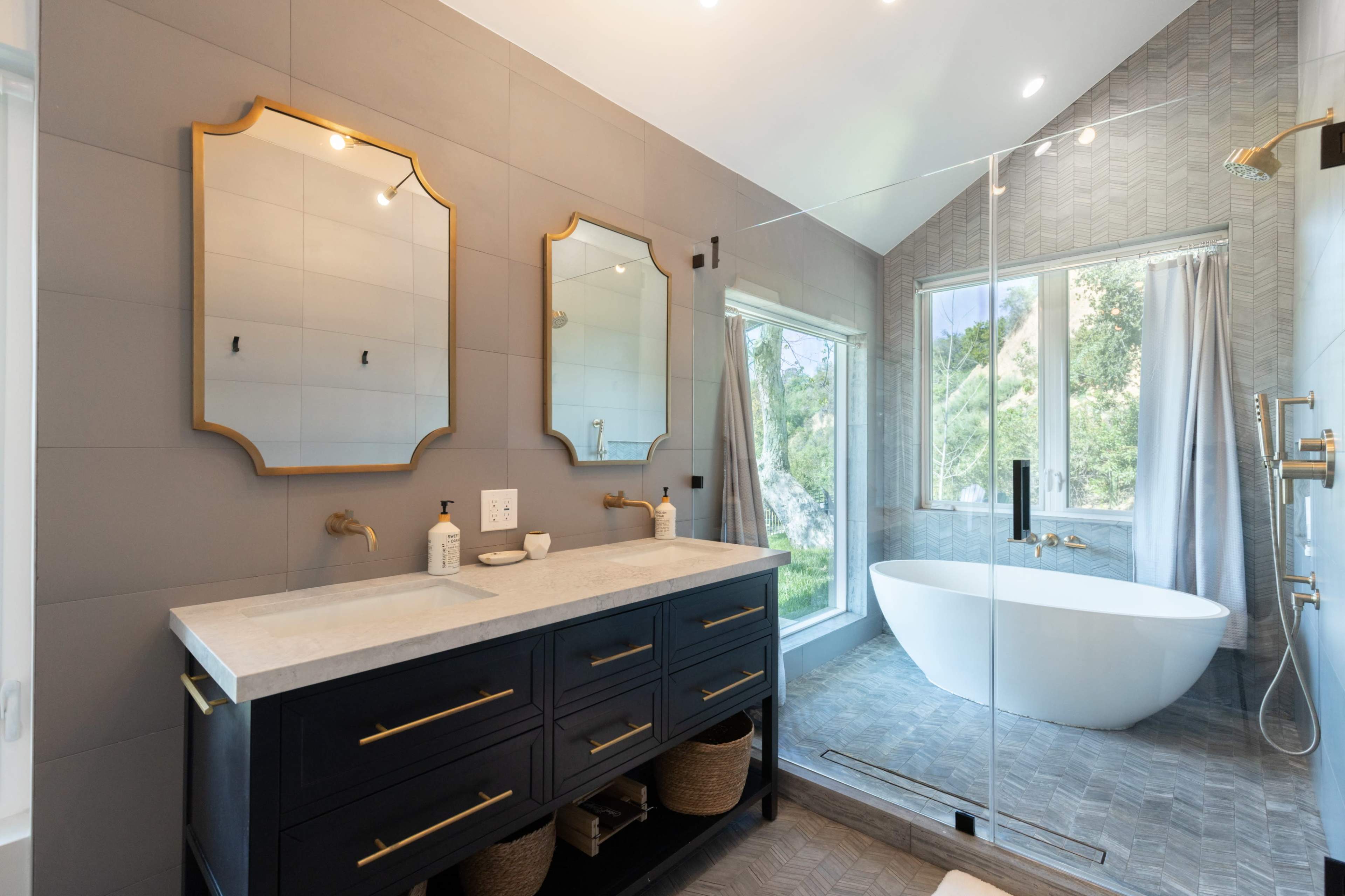 A modern bathroom features a double sink vanity with mirrors, a freestanding bathtub, and large windows overlooking greenery.