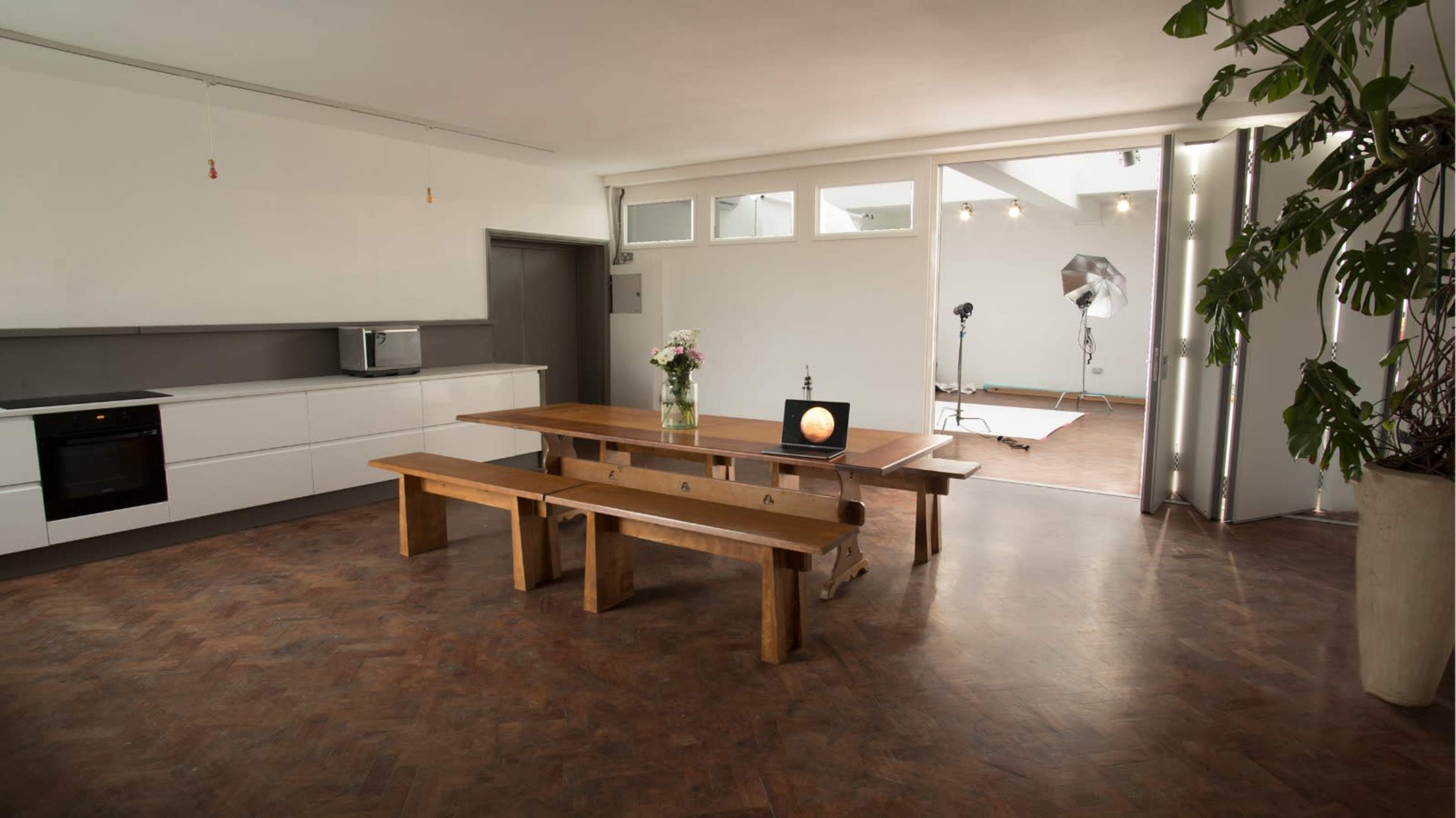 The image shows a modern, minimalist room with a kitchen area, a long wooden table, and a photo shoot setup in the background.