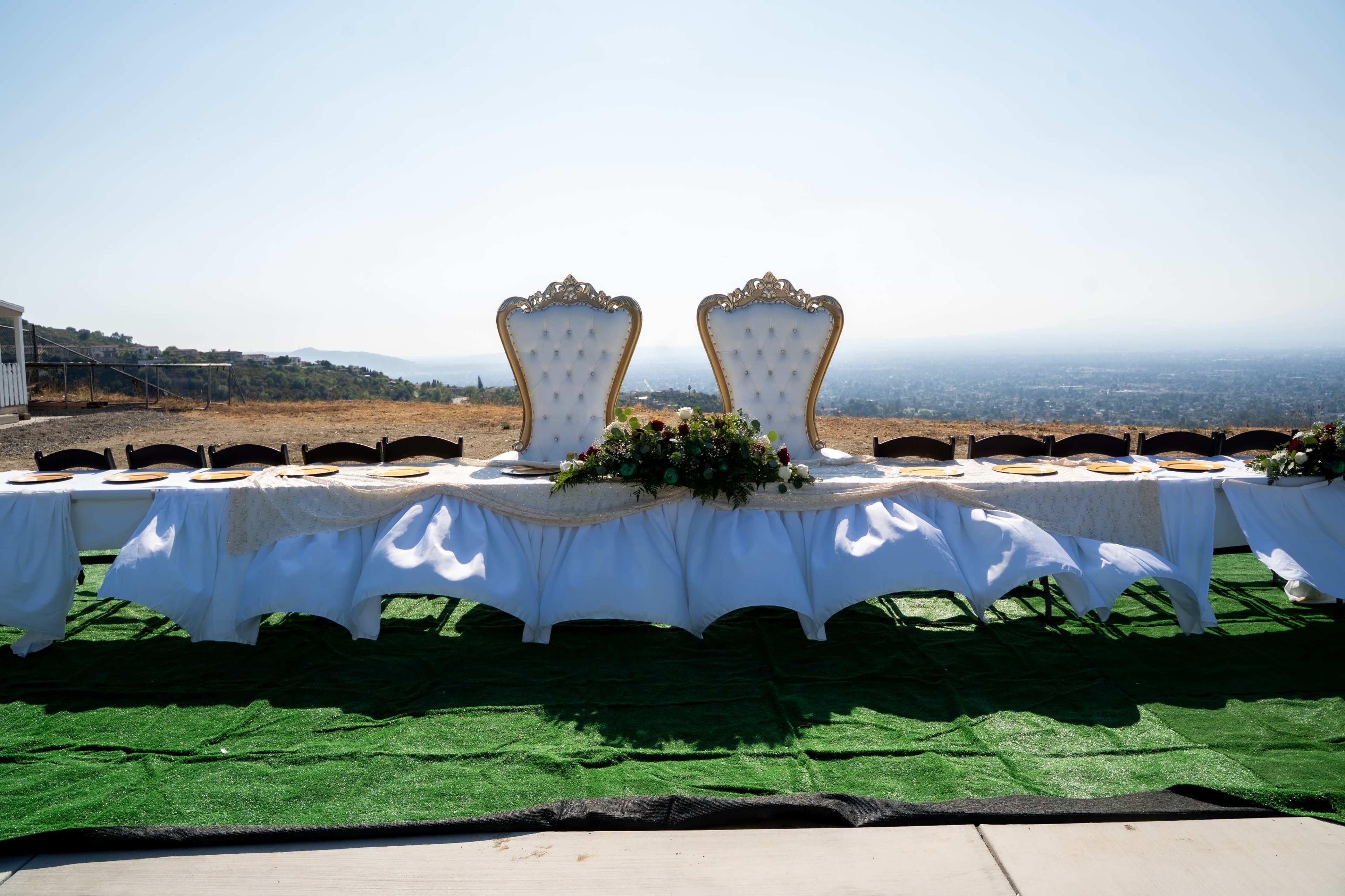 A long banquet table with two ornate chairs at the end is set on a grassy area, overlooking a distant landscape under a clear sky.