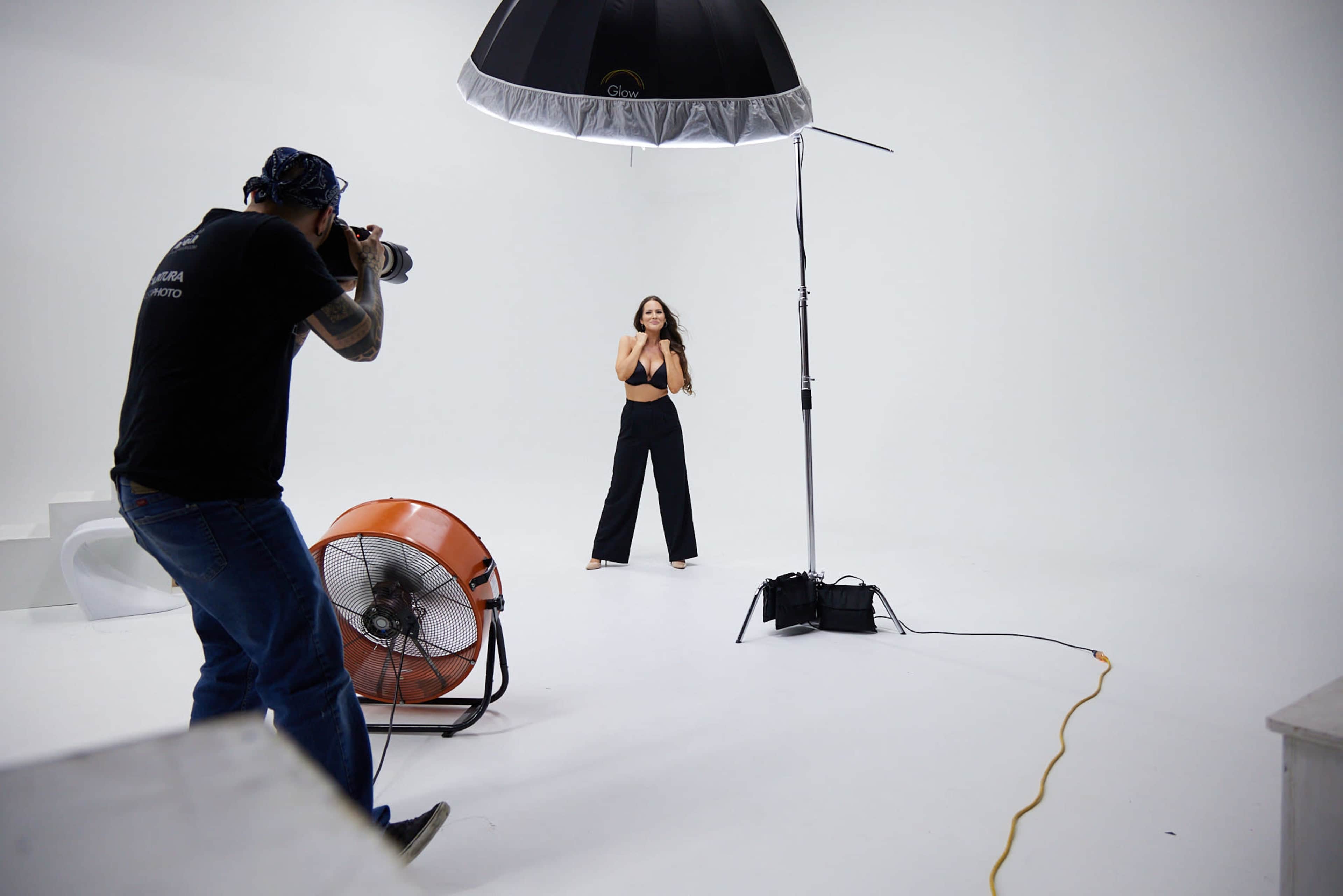 A photographer captures a model in a studio with bright white walls and a large softbox light overhead.