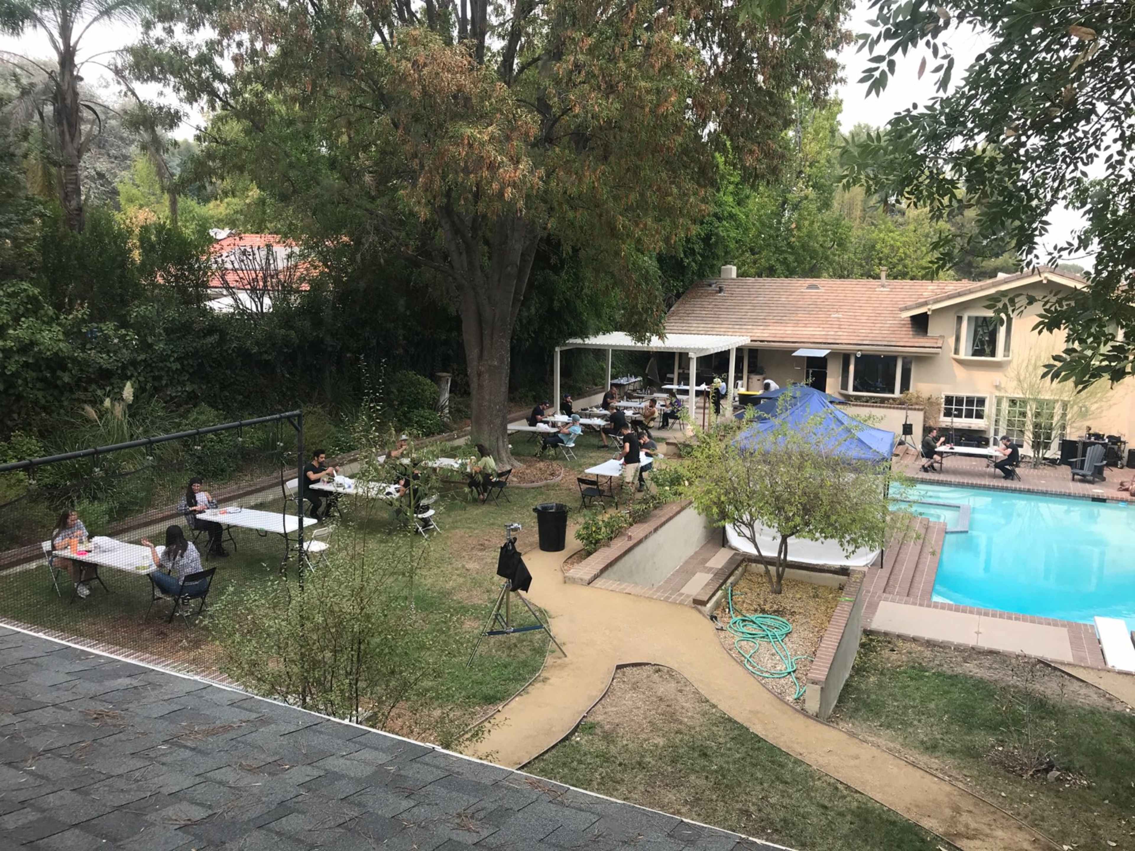 A group of people is seated at tables in a backyard near a swimming pool, surrounded by trees and a house in the background.