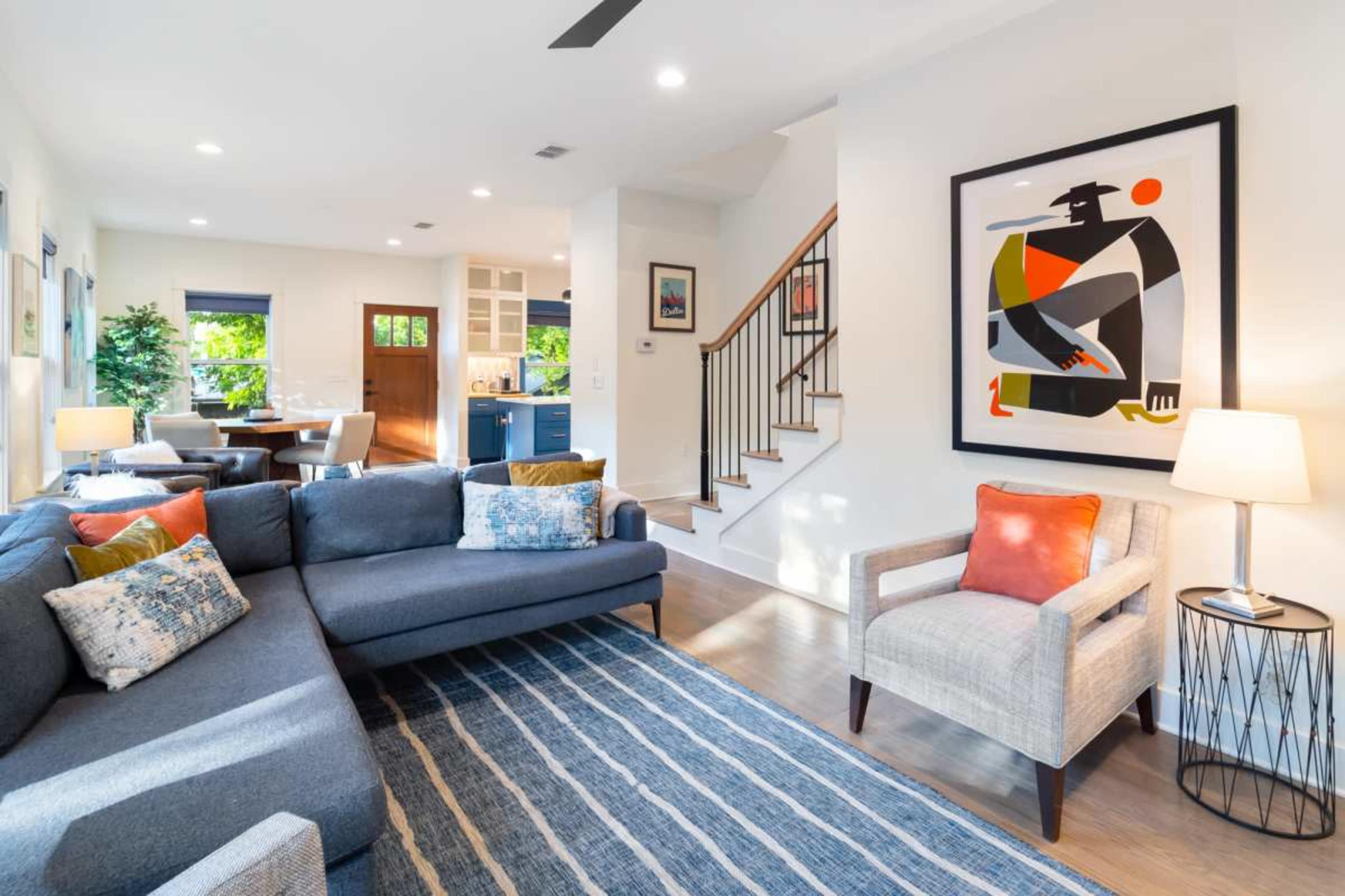 The image shows a modern living room with a gray sectional sofa, a light-colored armchair, an abstract artwork on the wall, a staircase, and a dining area visible in the background.