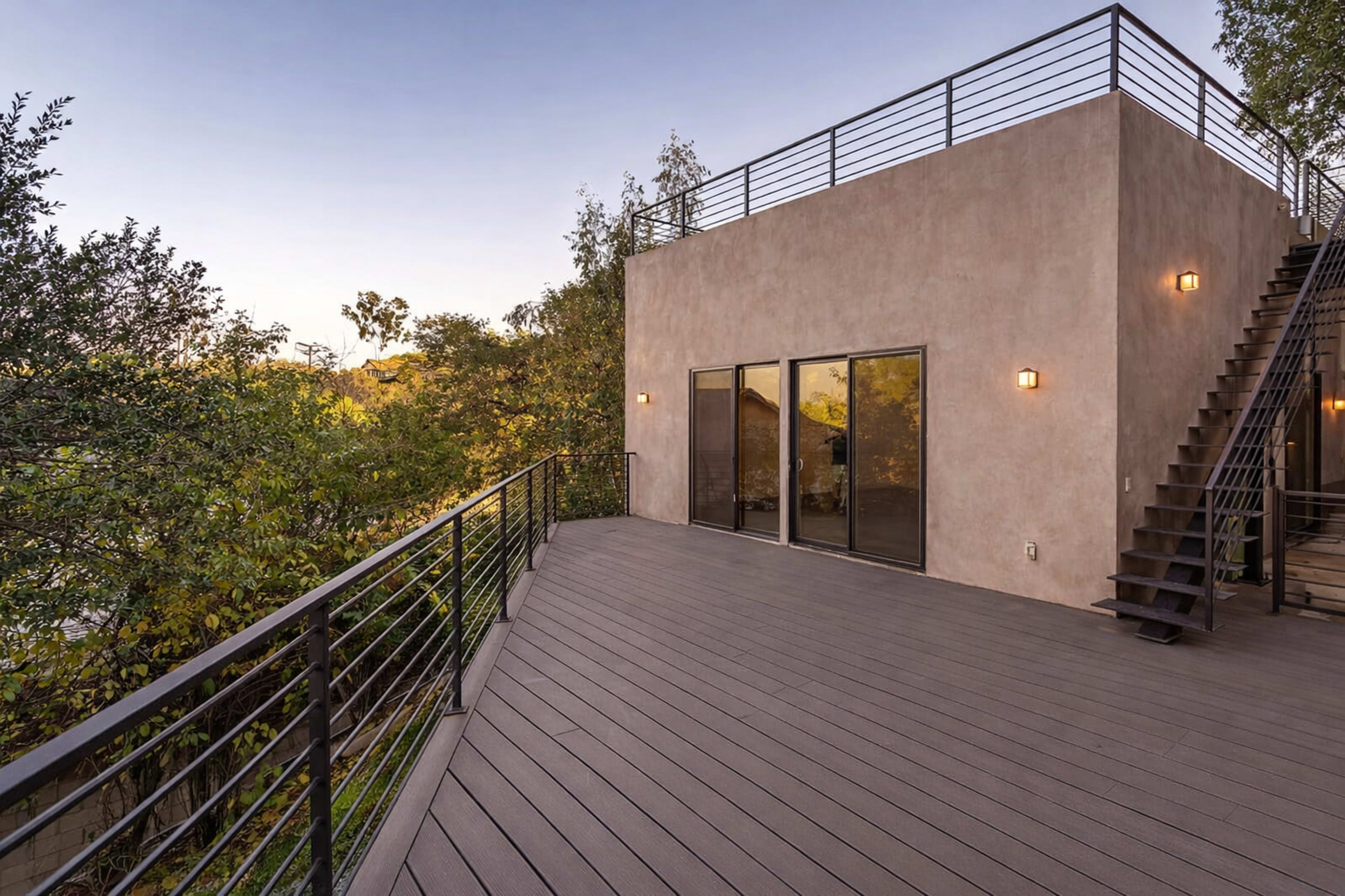 The image shows a modern deck with a view of trees and a building with large glass doors and exterior stairs.