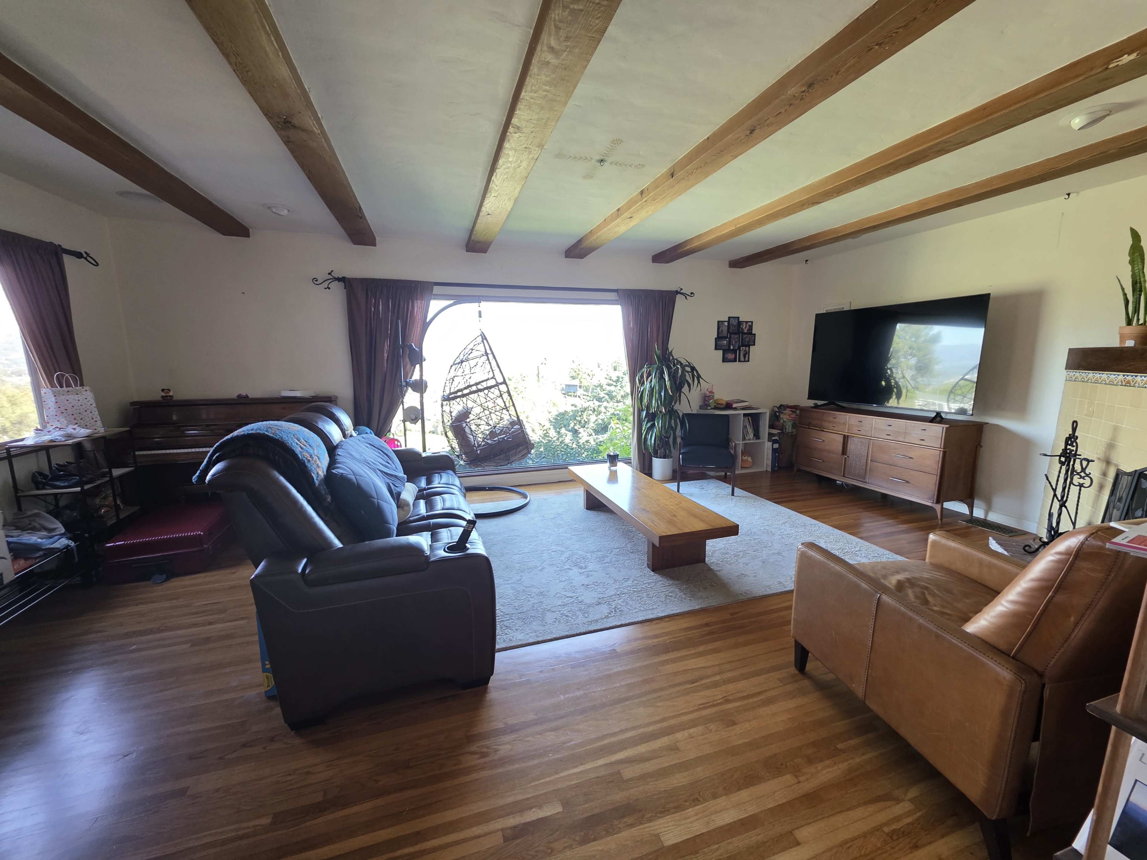 The image shows a spacious living room with wood beam ceilings, a large window offering a view, and various seating arrangements including a leather couch, an armchair, and a coffee table.