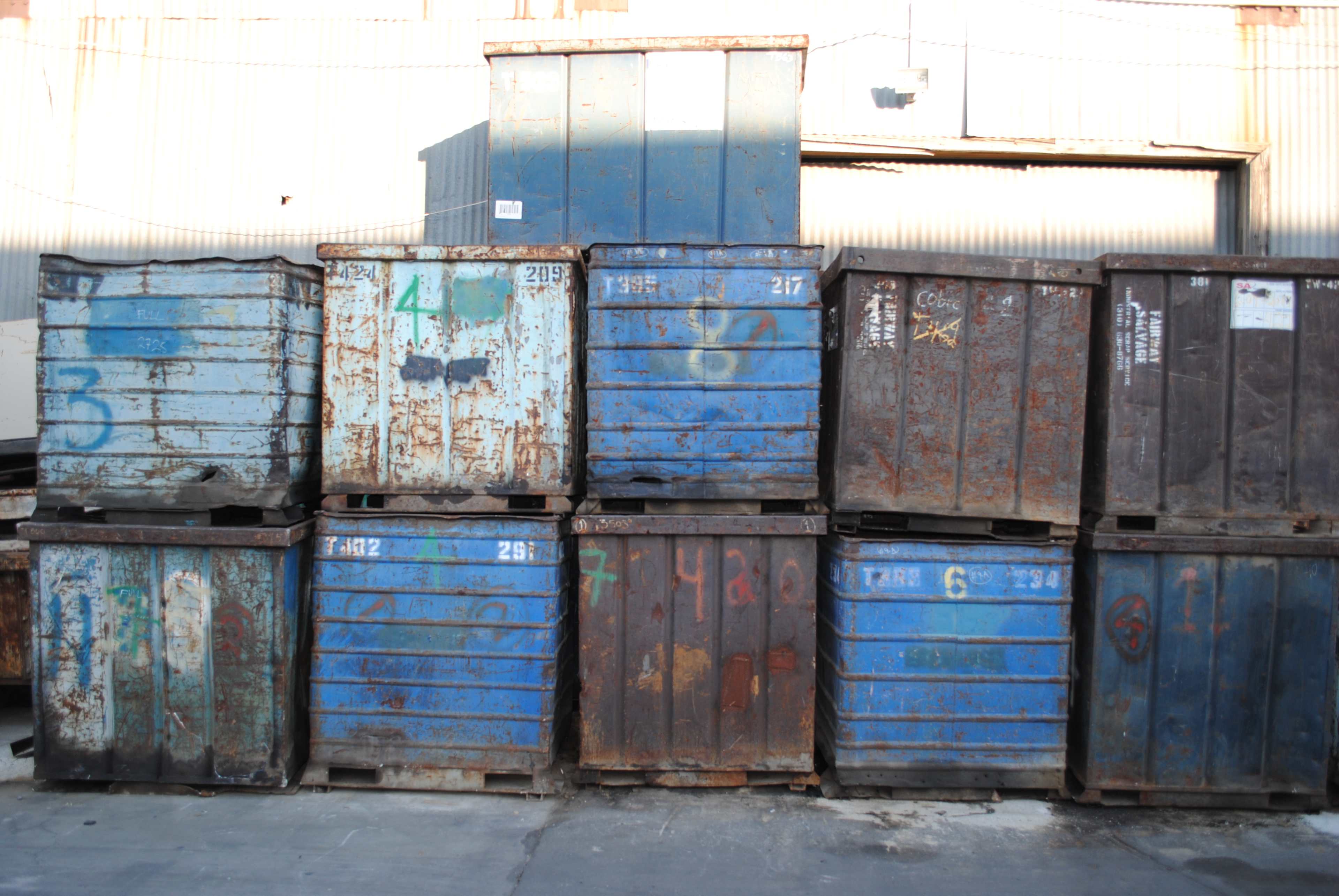 A stack of metal containers in various shades of blue, some with visible rust and graffiti, arranged against a textured wall.