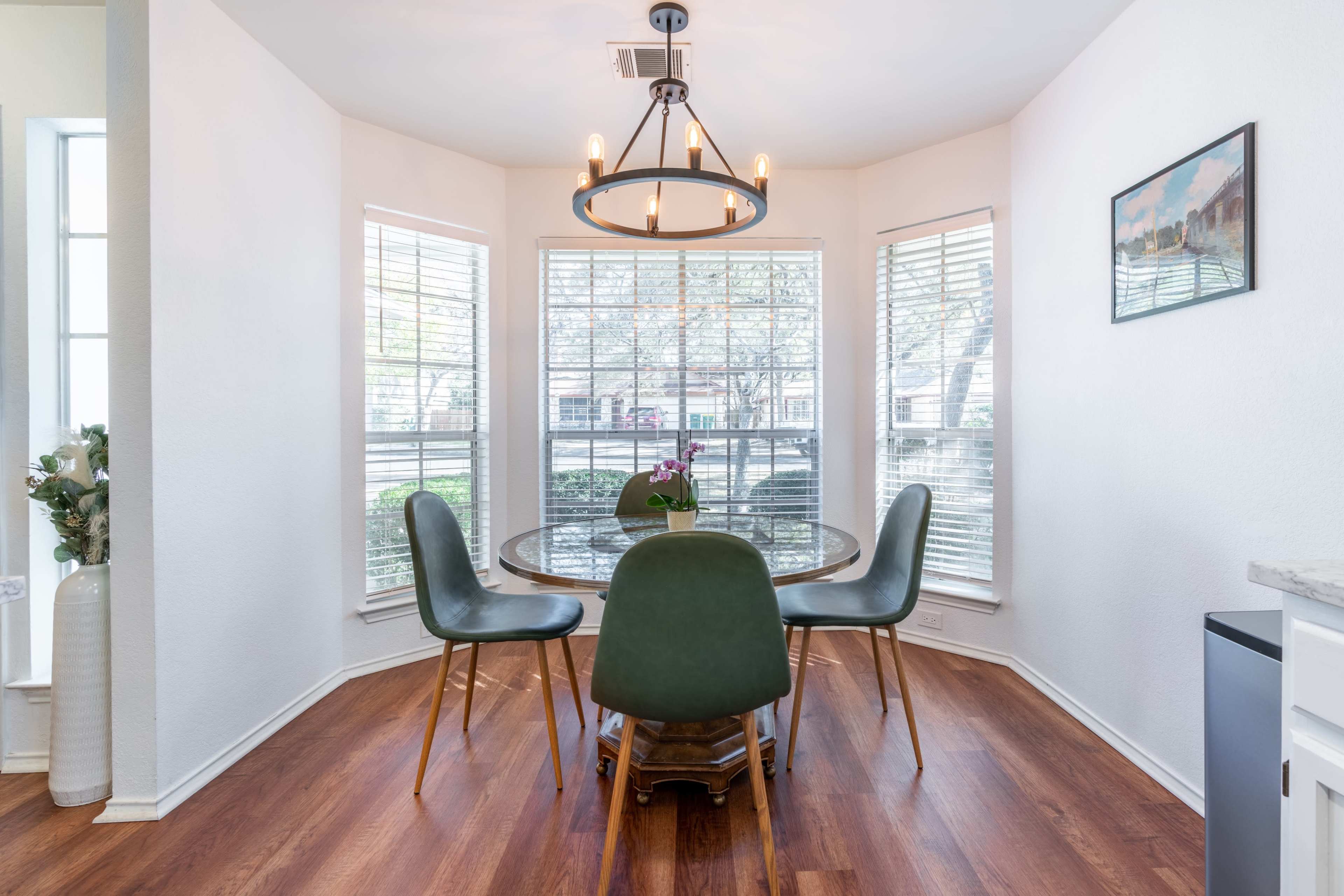 A circular glass dining table with green chairs is set in a sunlit corner surrounded by large windows.