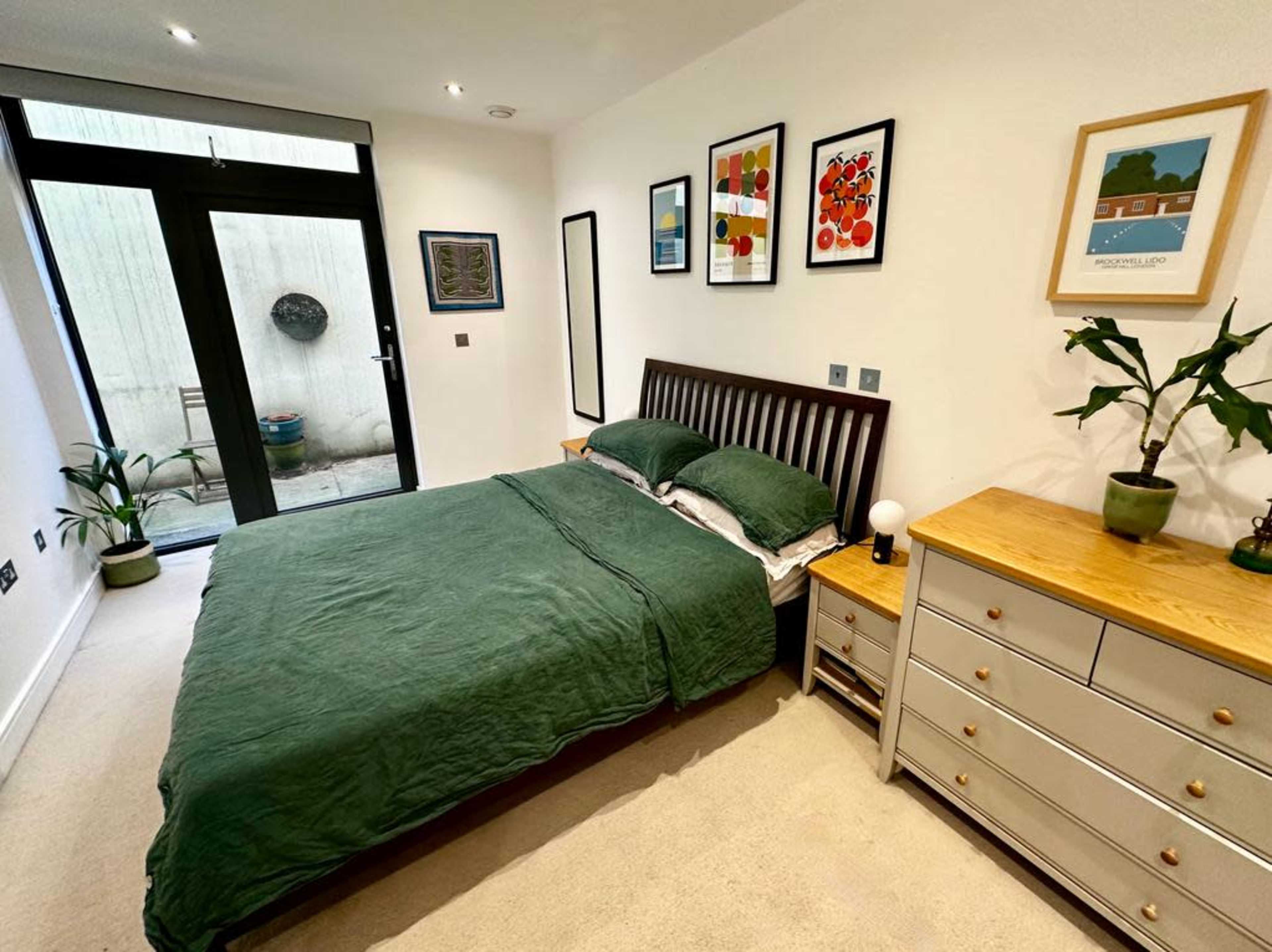 A bedroom with a green bedspread, a wooden bed frame, a dresser, and framed artwork on the walls, along with a sliding glass door leading to an outdoor space.
