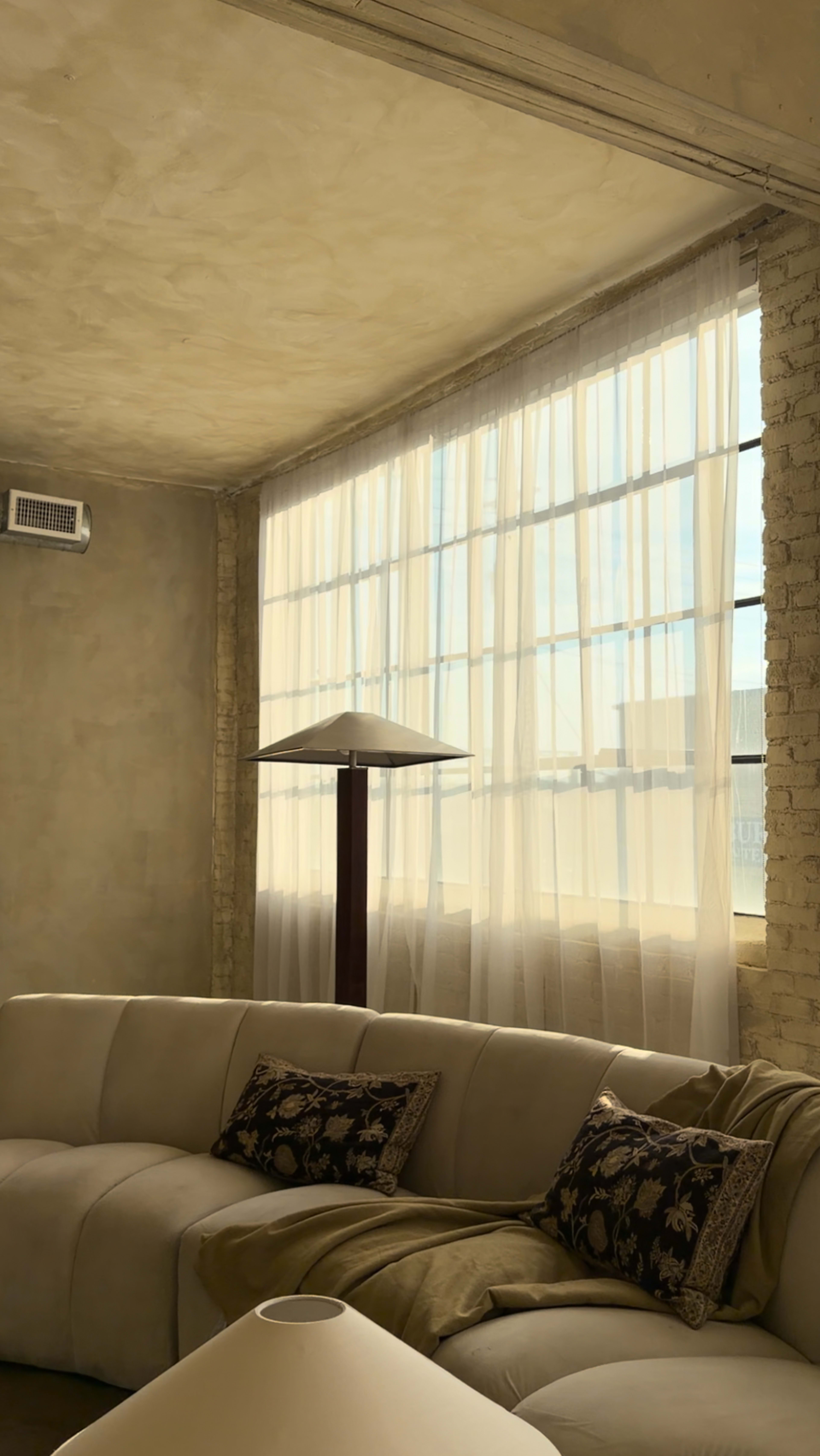 A soft, cream-colored couch with decorative pillows is positioned beside large, sunlit windows adorned with sheer curtains in a minimally furnished room.