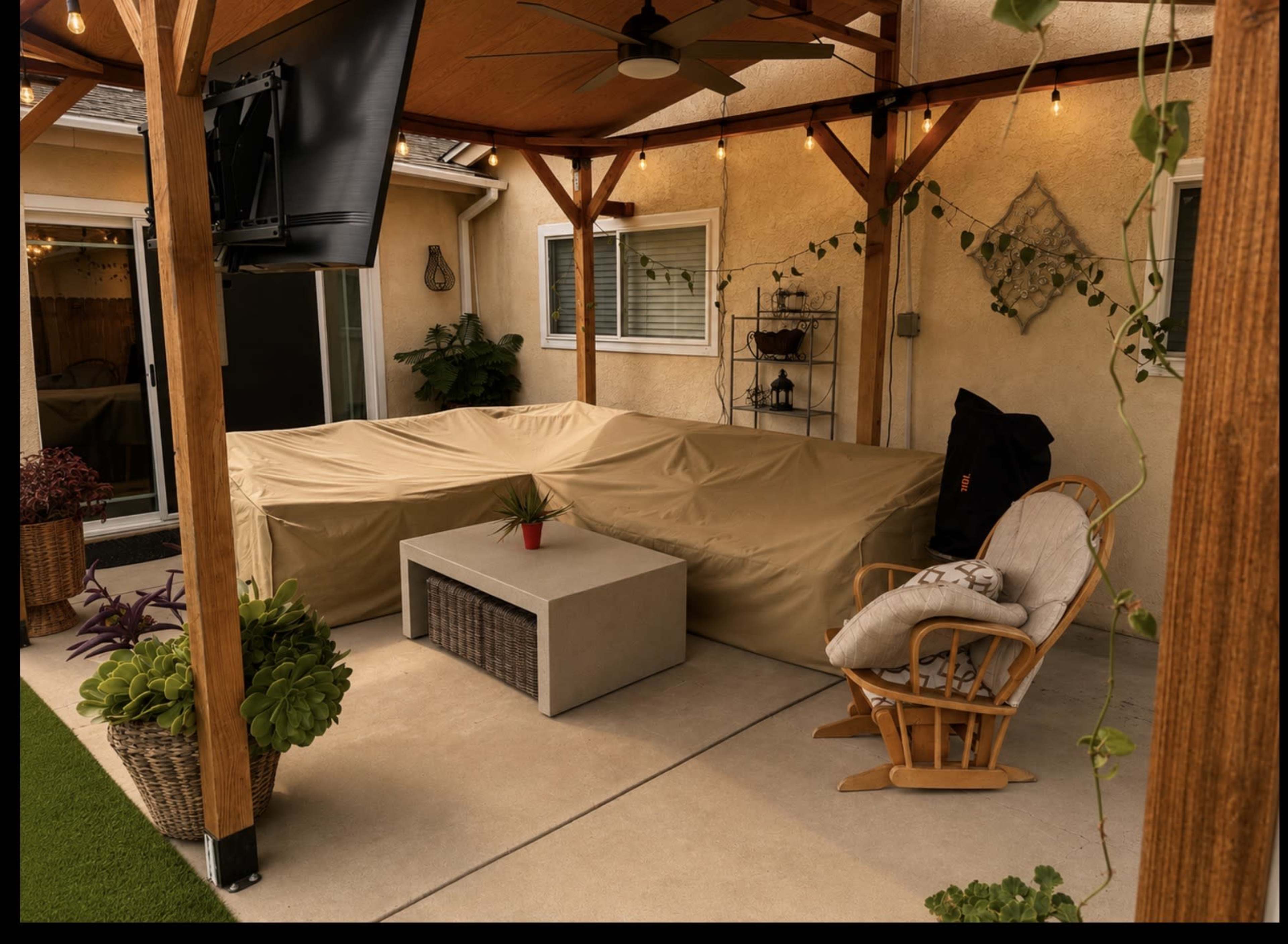 The image shows a covered outdoor seating area featuring a large, beige cover over a sectional sofa, a small coffee table, and a rocking chair under a wooden pergola with string lights.