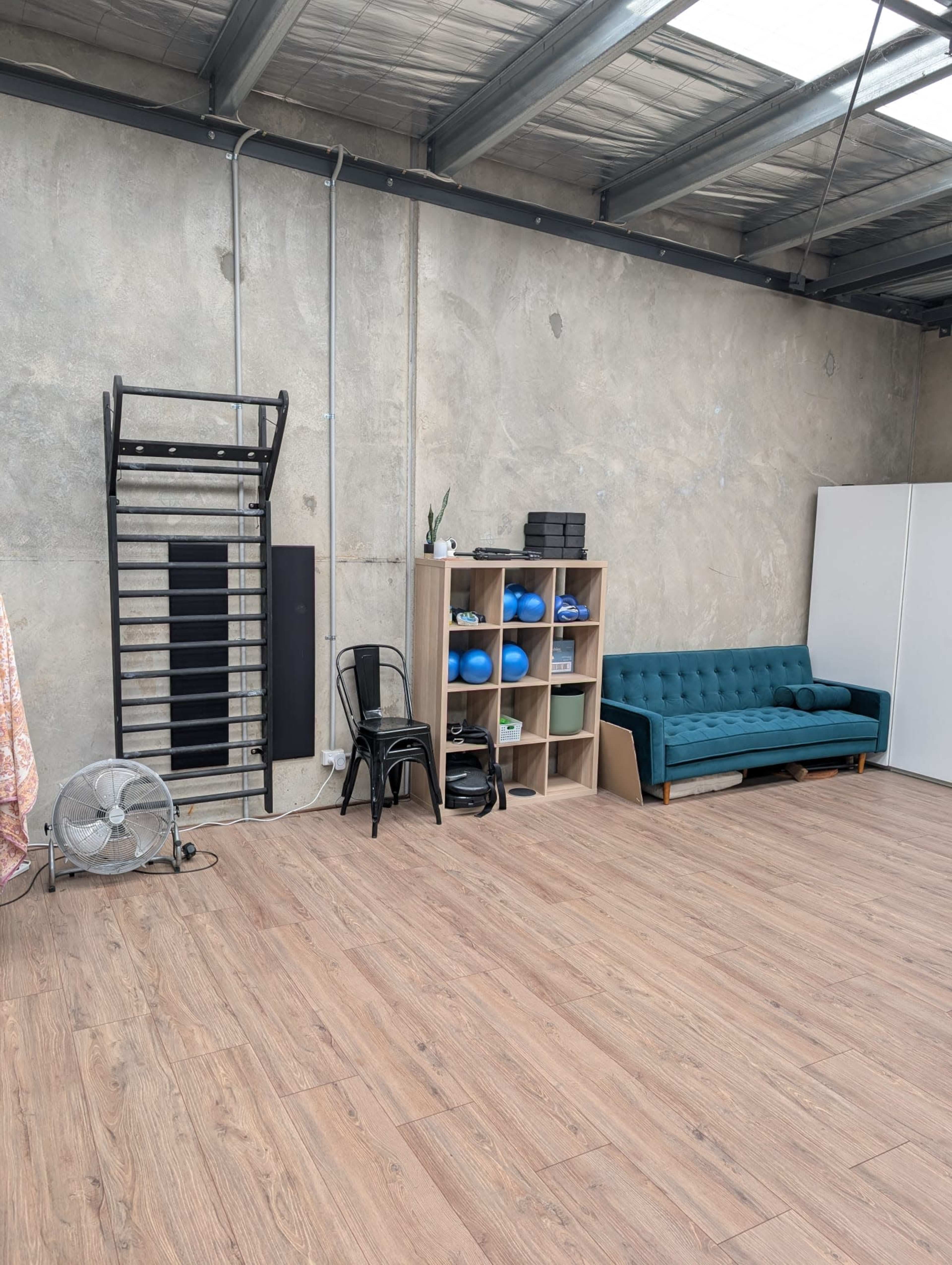 The image shows a spacious studio with a wooden floor, a green couch, a storage unit with various items, a fan, and a wall-mounted exercise bar.