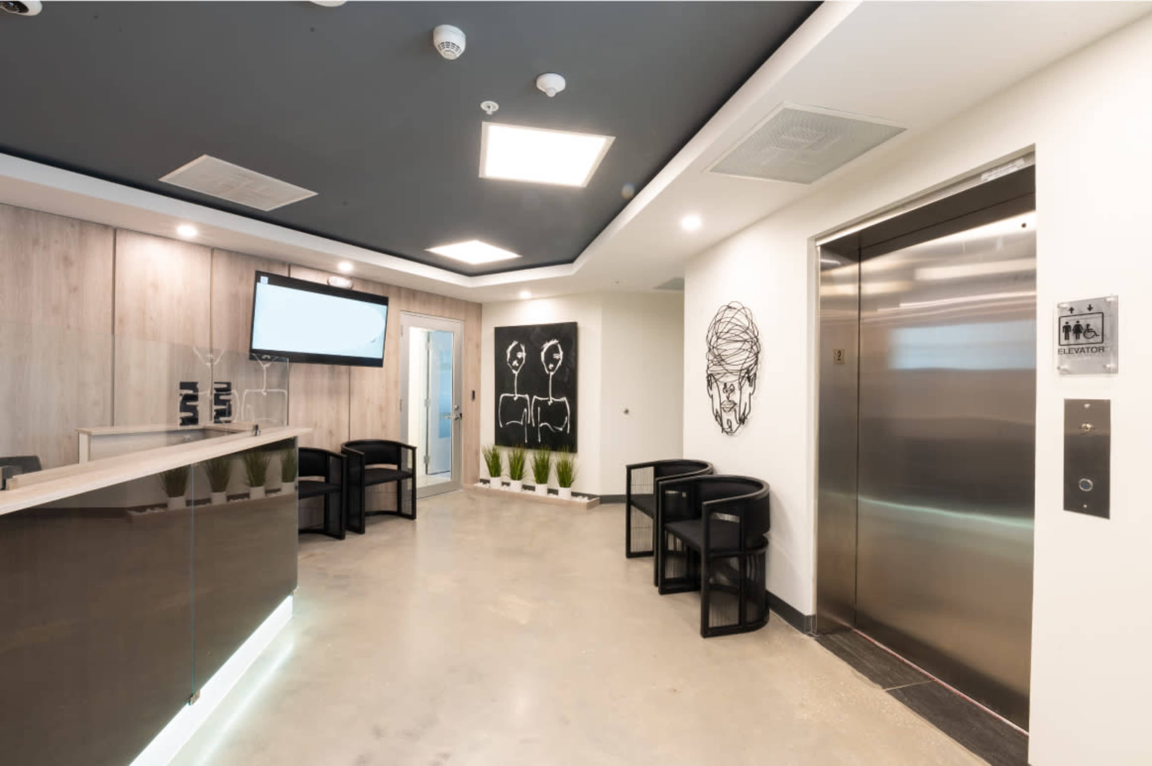 A modern reception area with a sleek front desk, an elevator, black and white wall art, and several chairs arranged near the entrance.