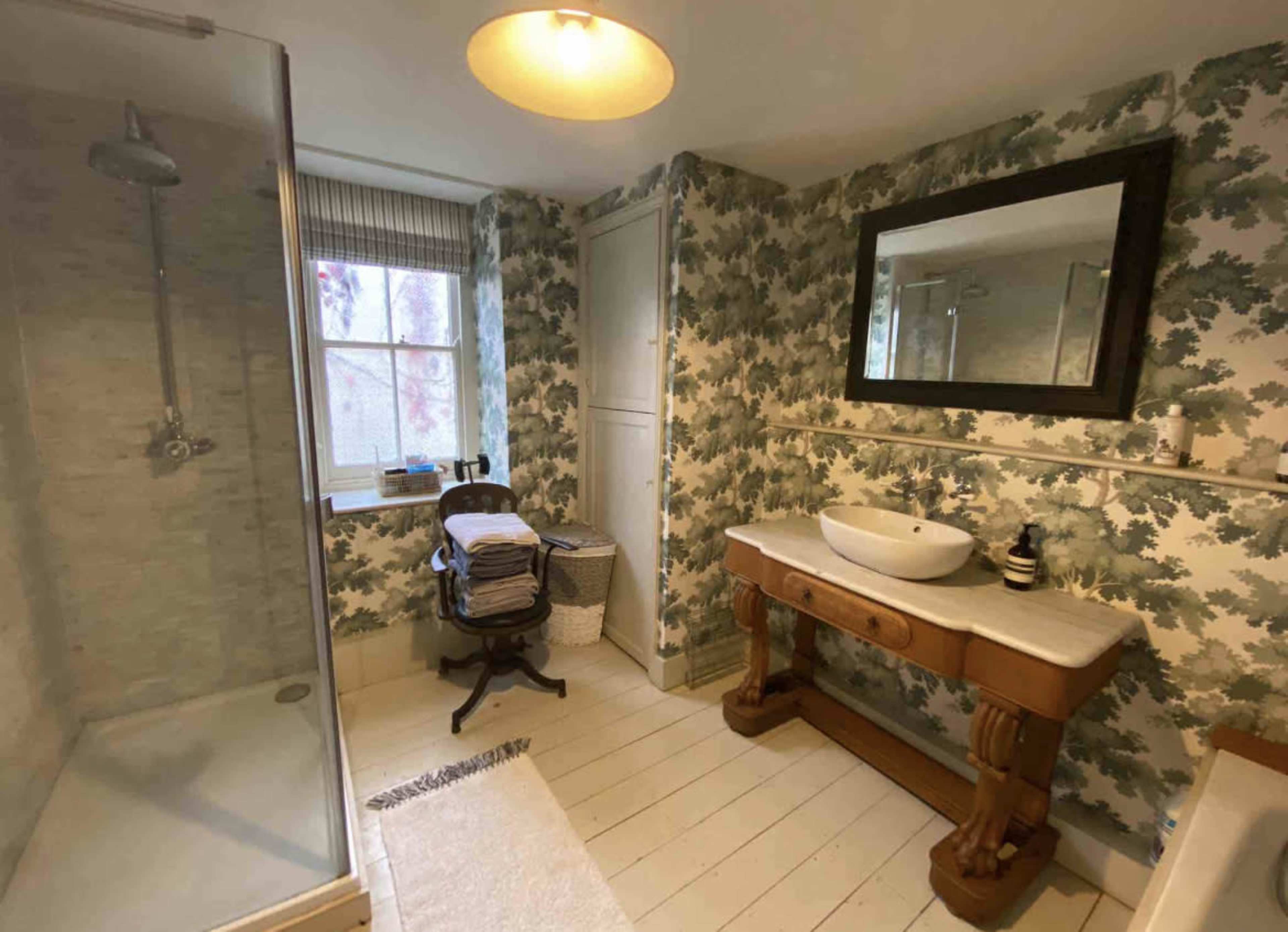 A bathroom with a glass shower, a wooden vanity with a sink, stacks of towels on a chair, and patterned wallpaper.