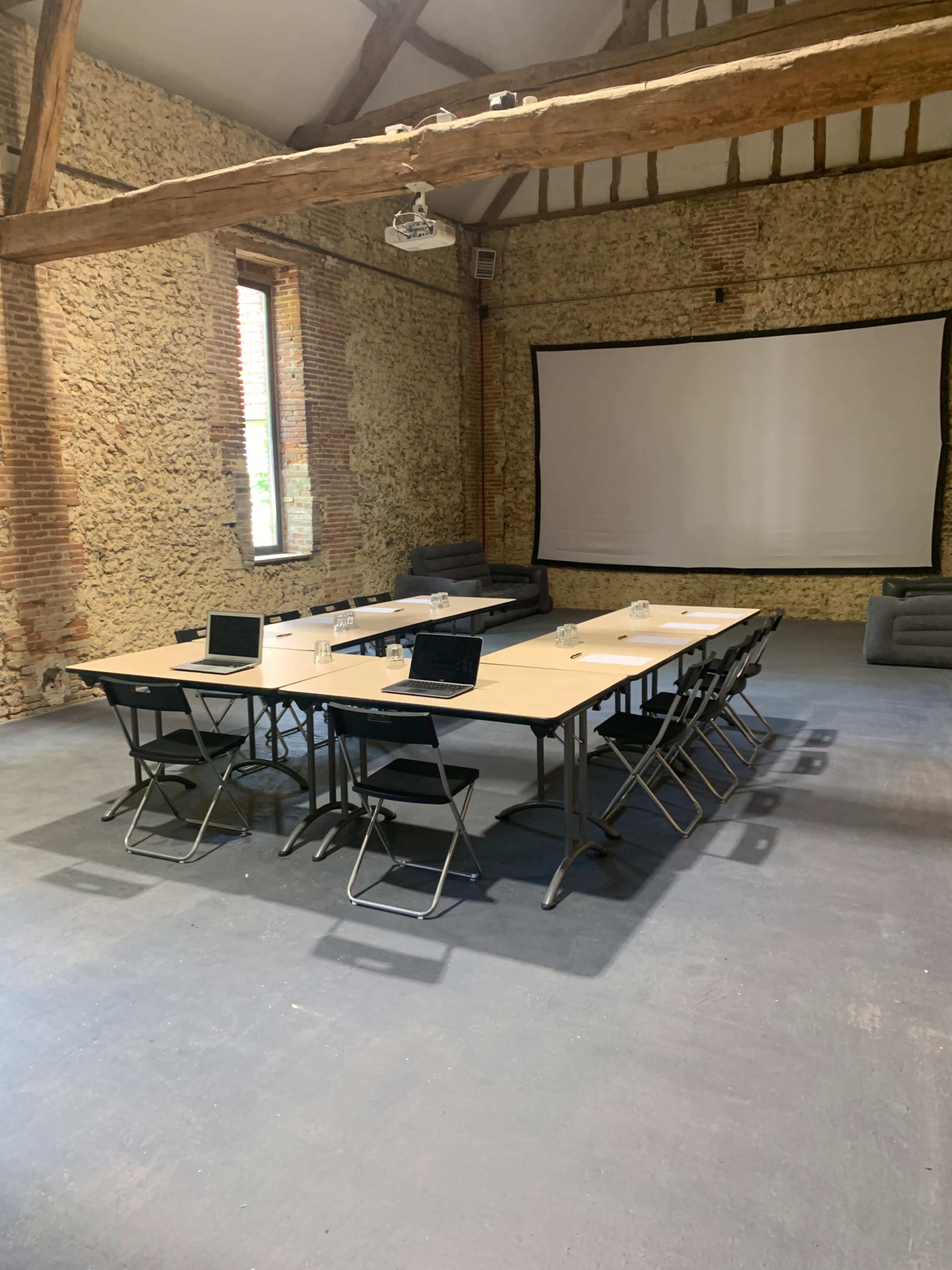 A spacious meeting room features a long table with laptops and glassware, surrounded by folding chairs, and a large screen mounted on the wall.