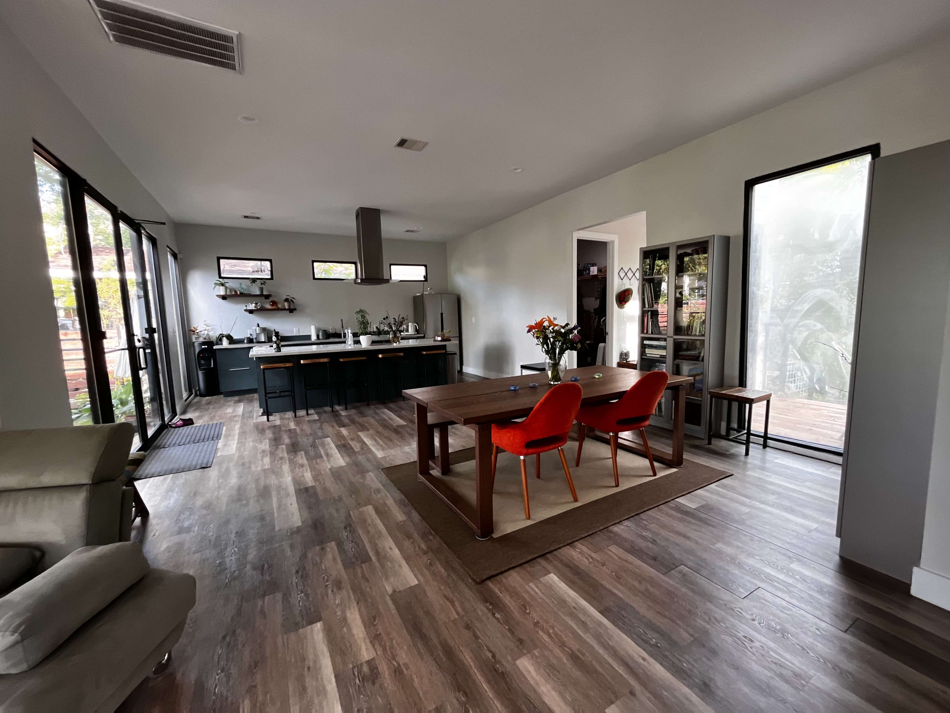 The image shows a modern open-concept living and dining area with a kitchen, a wooden dining table surrounded by red chairs, and large windows that provide natural light.