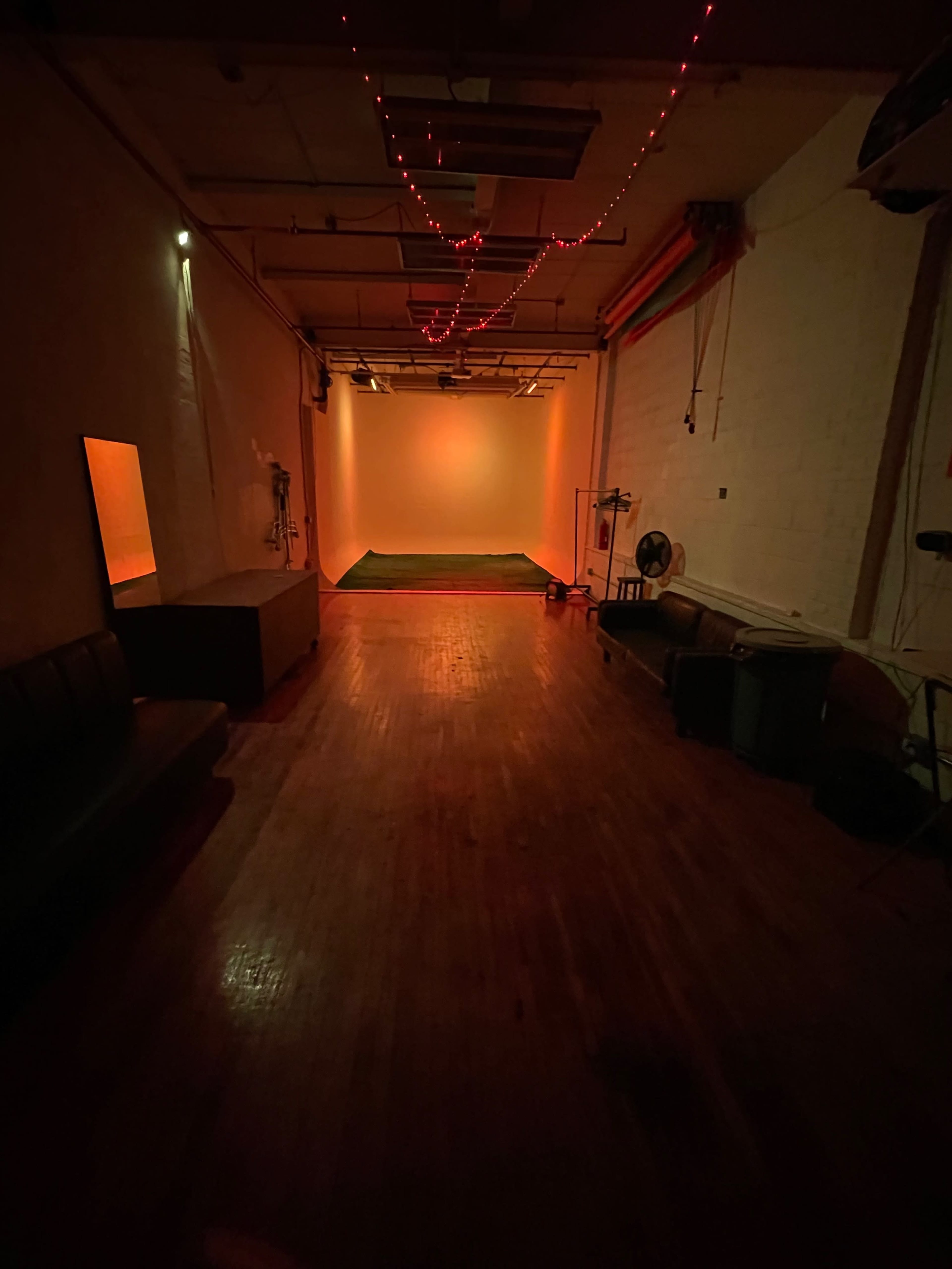 An illuminated studio space with a wooden floor, orange lighting at the back, and scattered furniture along the walls.
