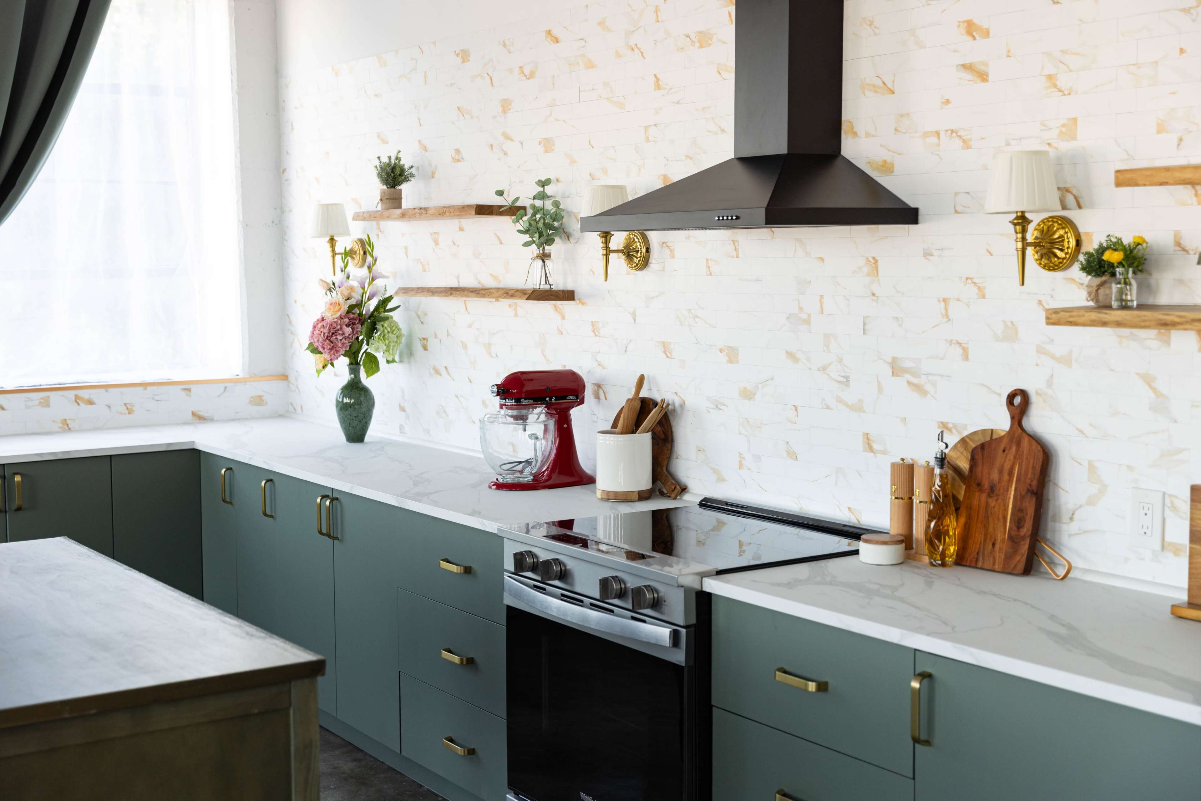 The image shows a modern kitchen featuring green cabinets, a stainless steel stove, and a red stand mixer on a countertop with white stone backsplash and wooden shelves displaying decorative plants.