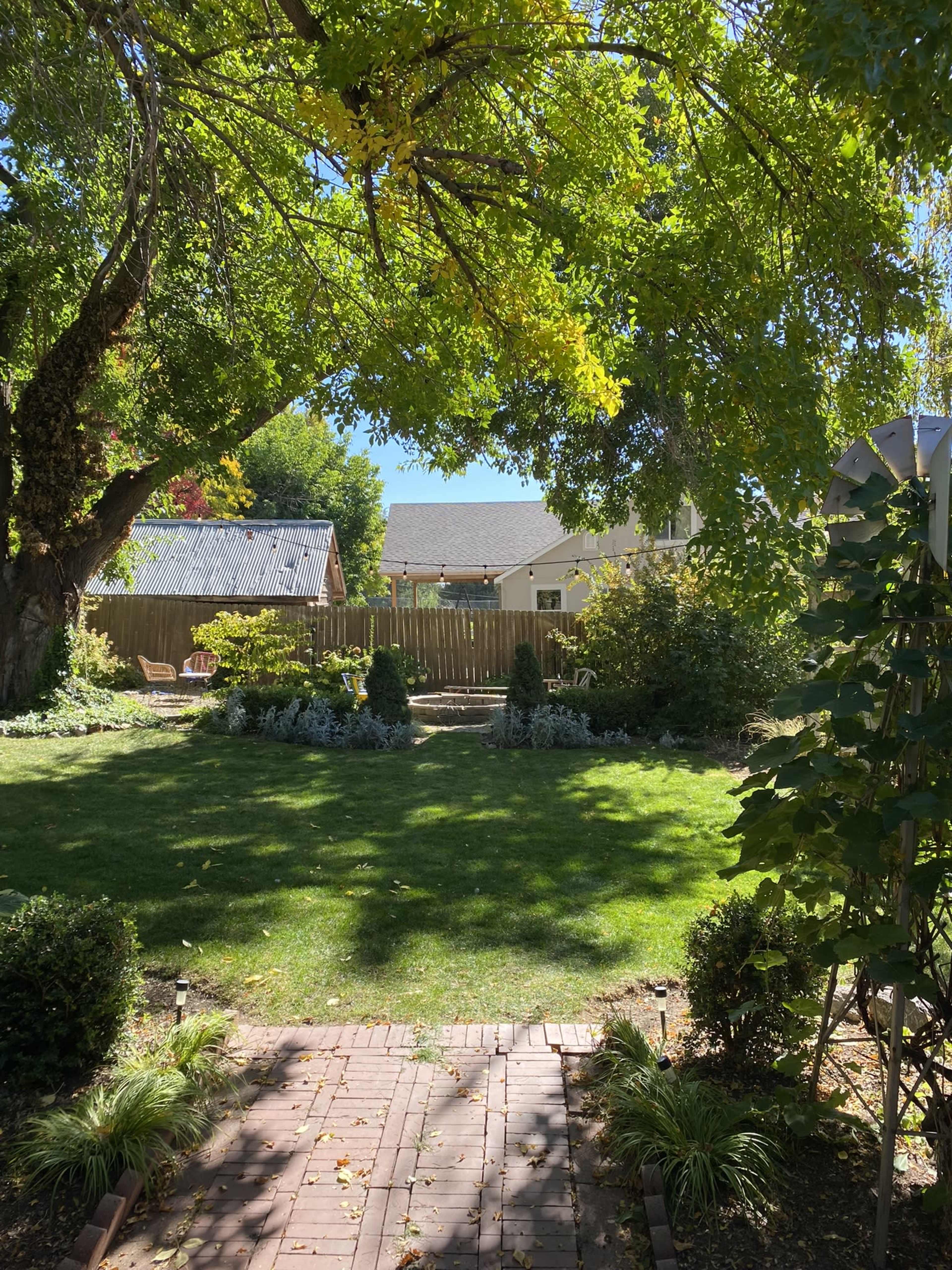 A brick pathway leads through a lush green yard surrounded by trees and shrubs, with a wooden fence and houses in the background.