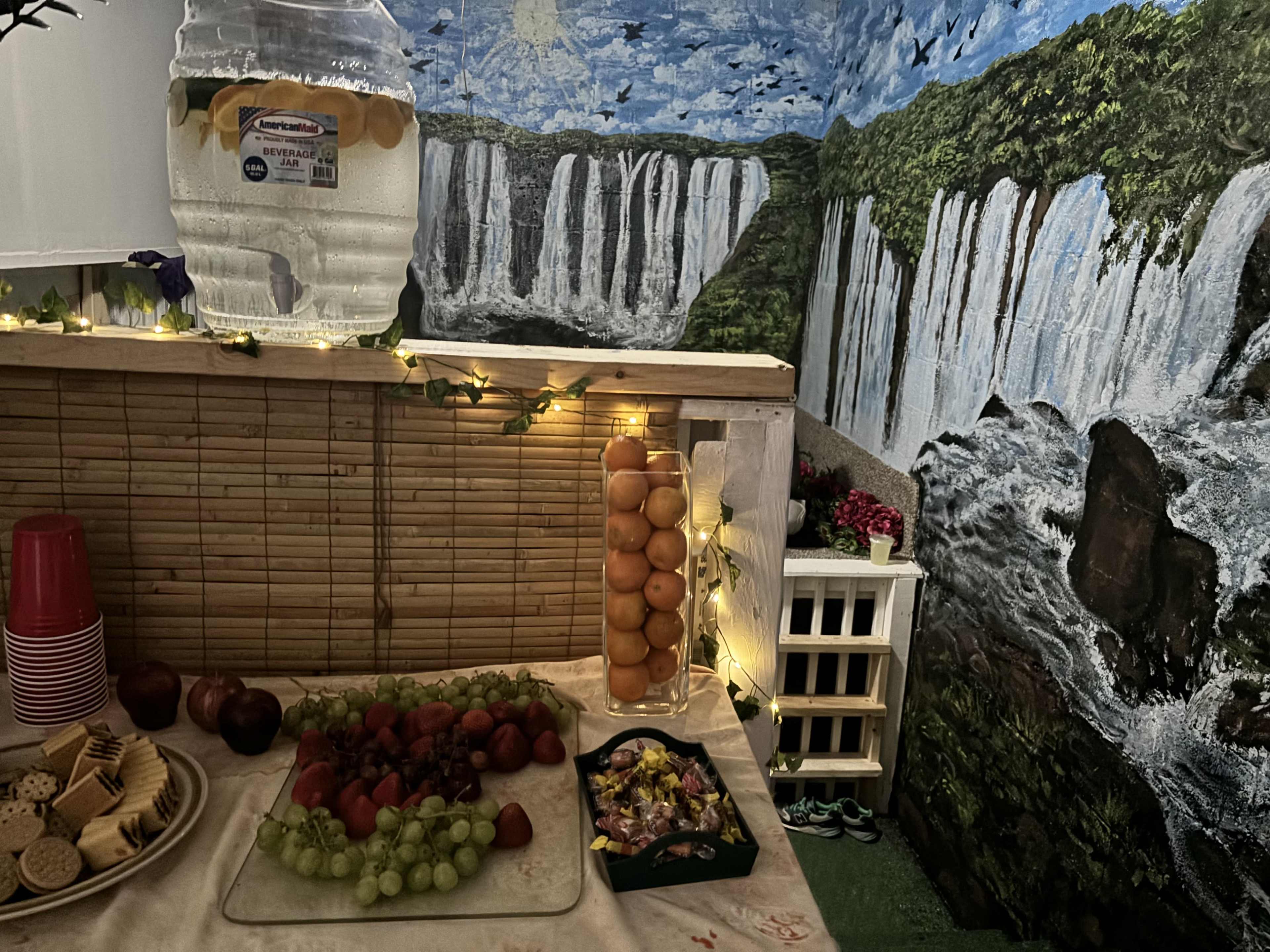 The image shows a decorated corner with a bamboo wall, a table featuring an assortment of fruits and snacks, and a vibrant mural of waterfalls in the background.