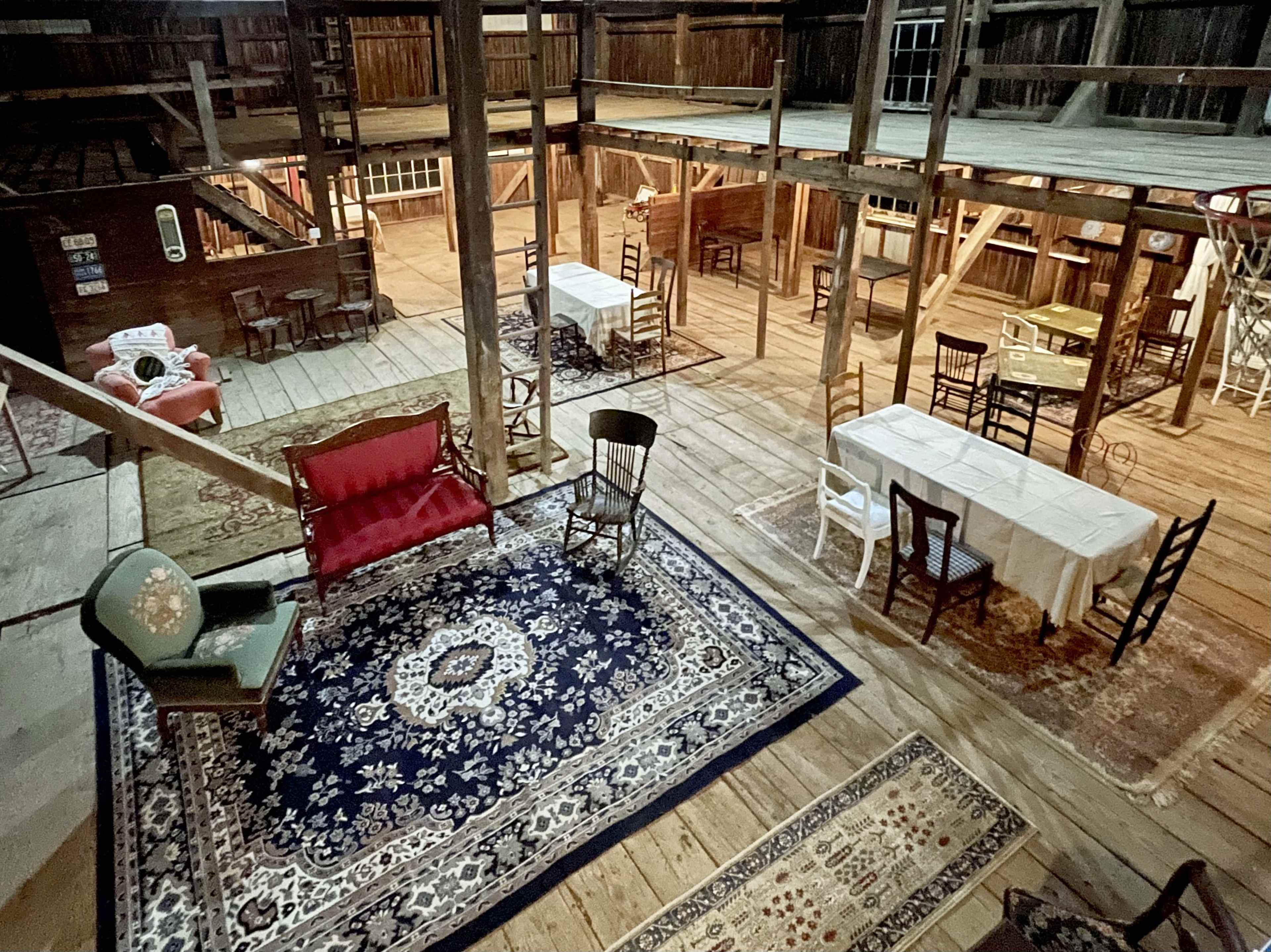 The image shows a spacious wooden interior with various tables and chairs arranged around a large rug, illuminated by overhead natural light.