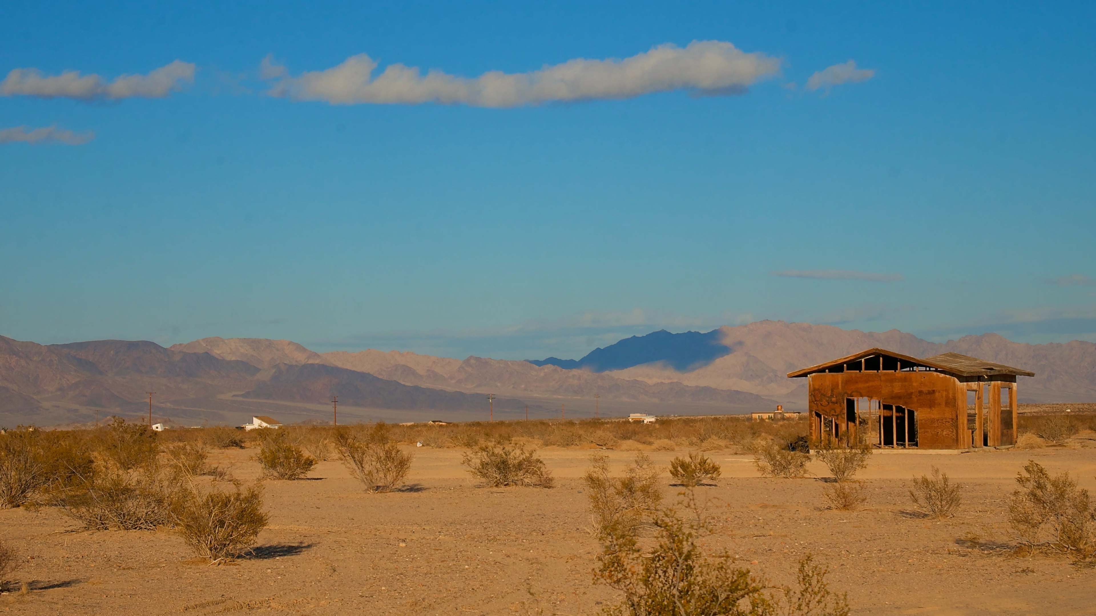 A rusty building stands in a barren landscape with sparse vegetation and mountains in the distance under a clear blue sky.