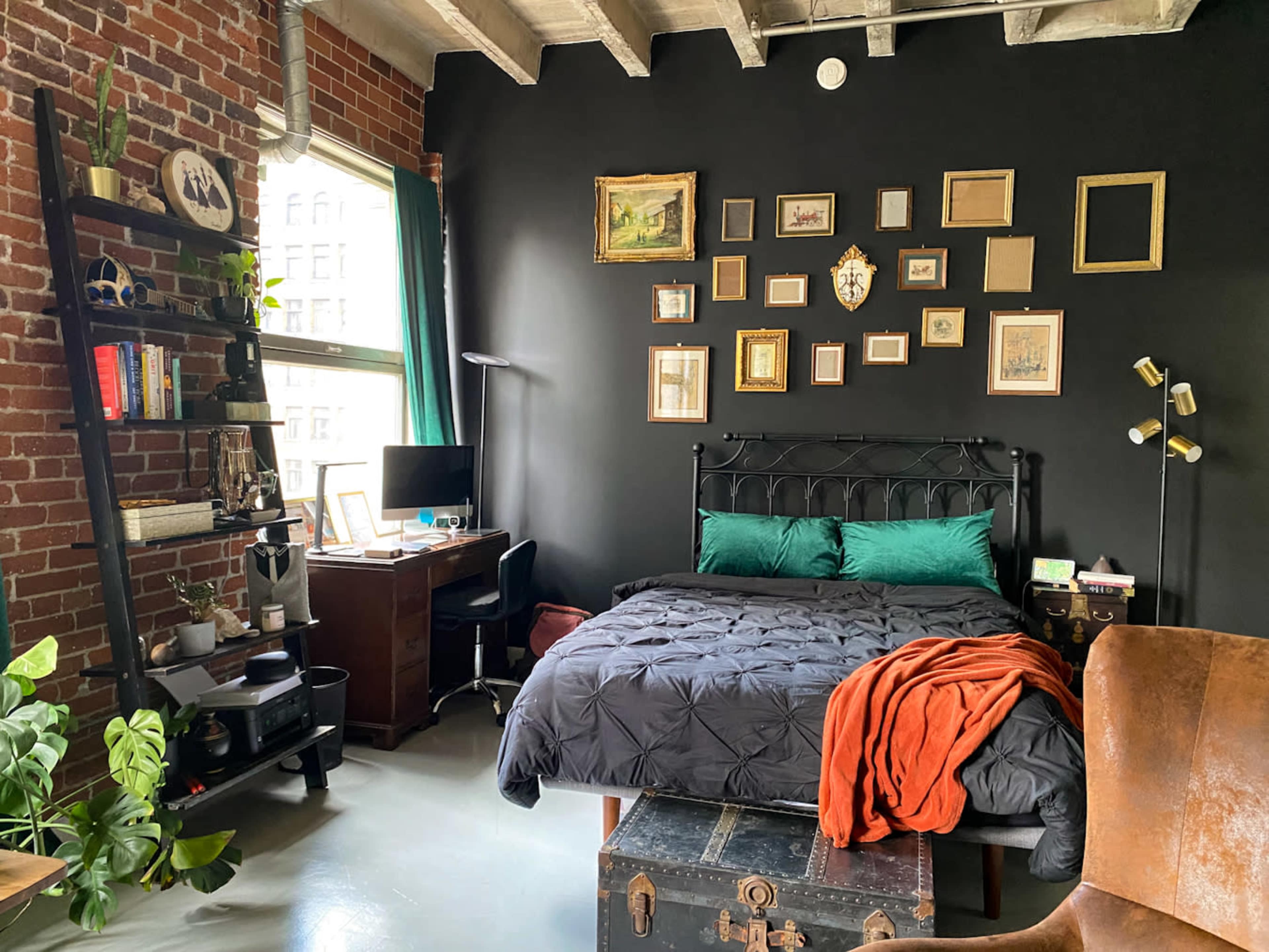 The room features a black wall behind a bed with green pillows, a bookshelf filled with various items, a desk with a lamp, and a mix of indoor plants and a vintage trunk.