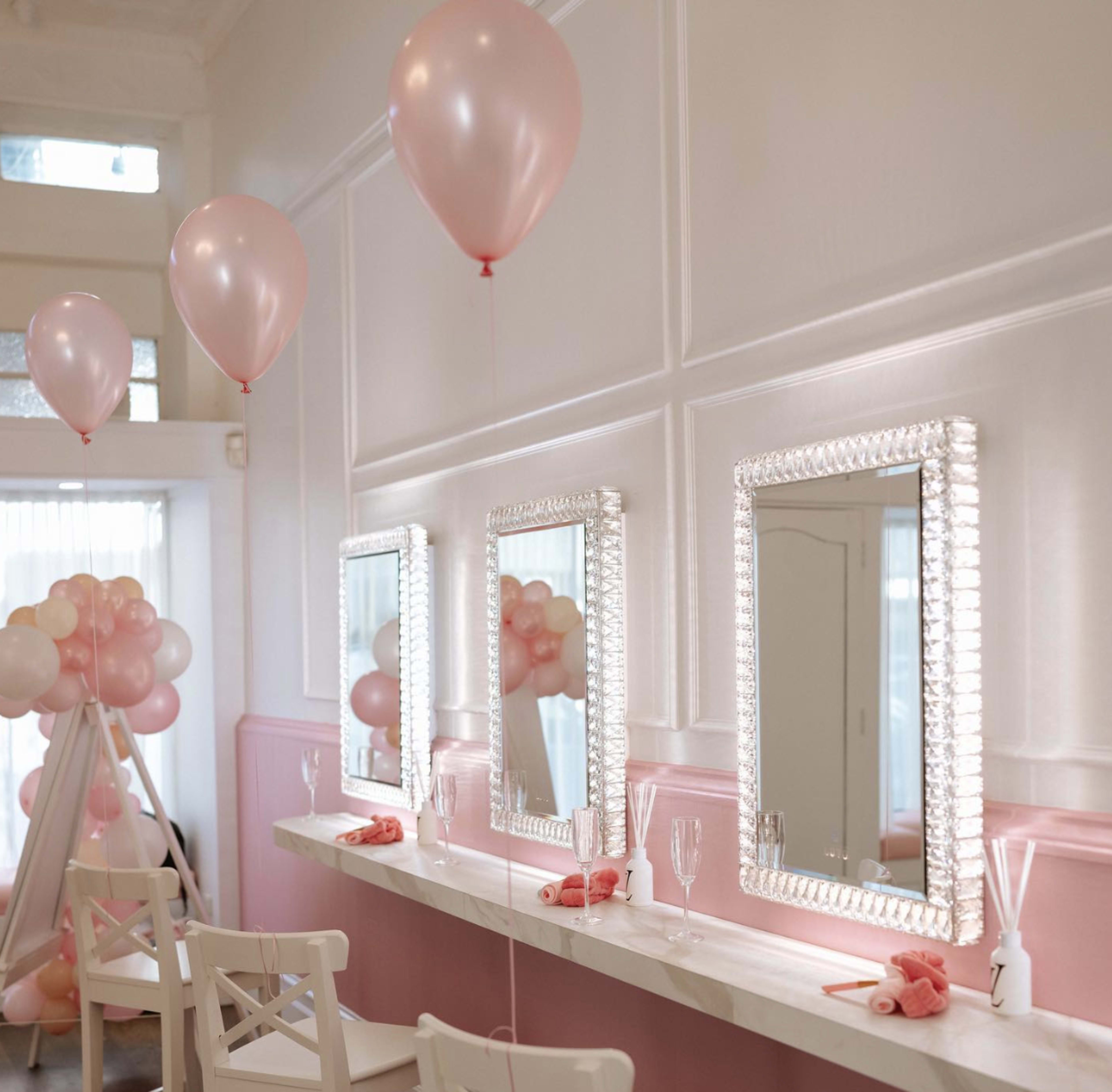 The scene features a brightly lit room with three decorative mirrors above a marble counter, pink balloons hanging from the ceiling, and a small display of balloons arranged to one side.