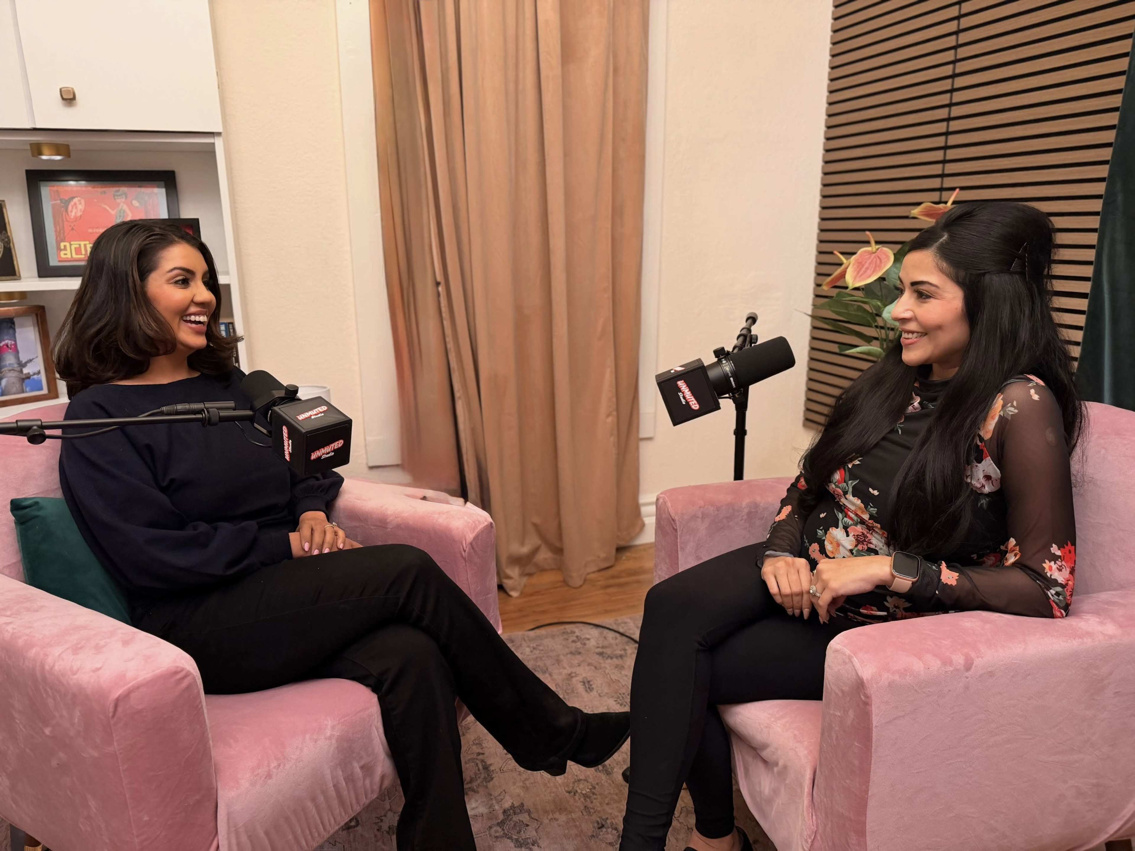 Two women are sitting in pink armchairs in a well-decorated room, engaging in conversation with microphones in front of them.