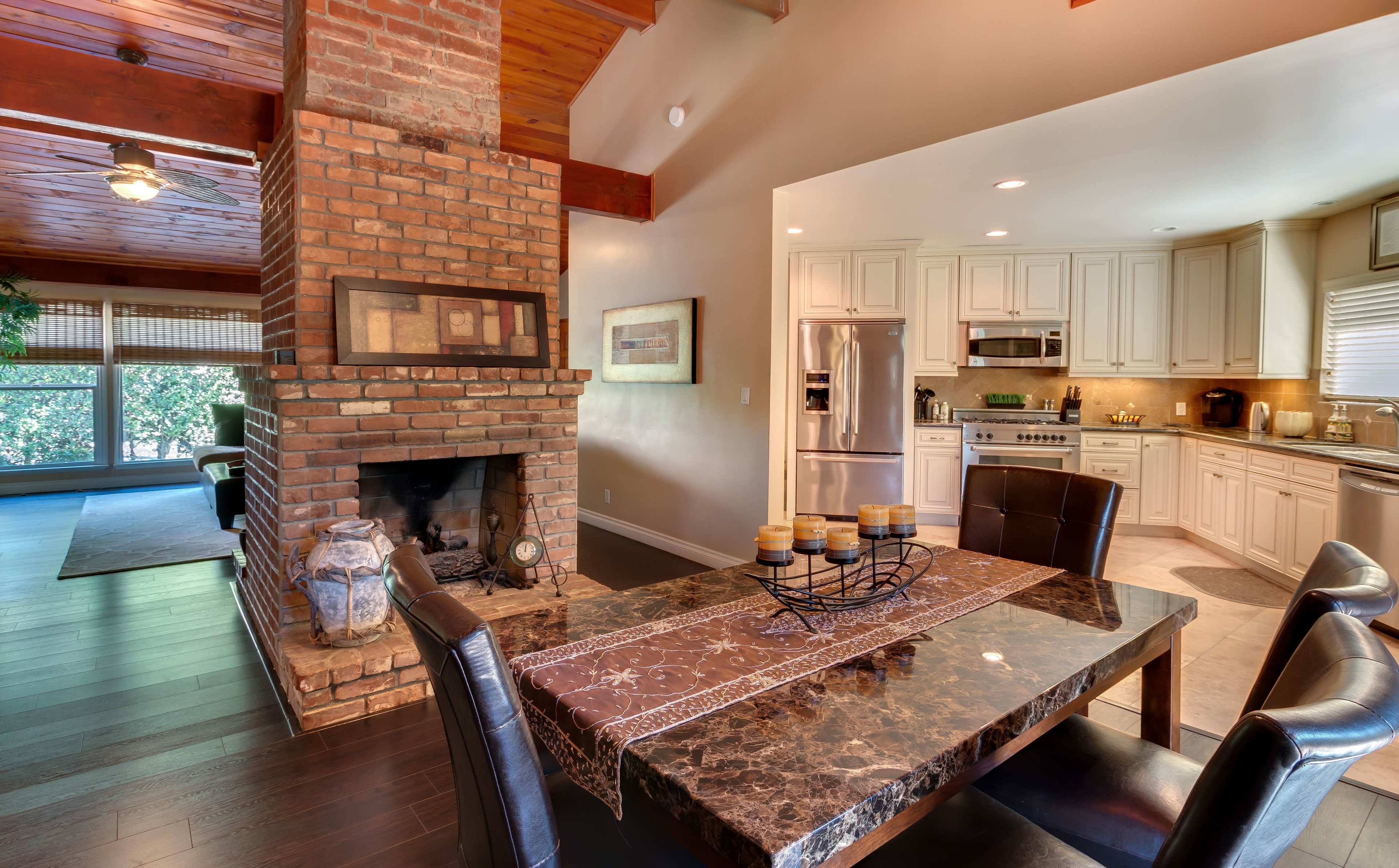 A spacious dining area with a dark marble table, a brick fireplace, and an adjoining kitchen featuring light-colored cabinets.