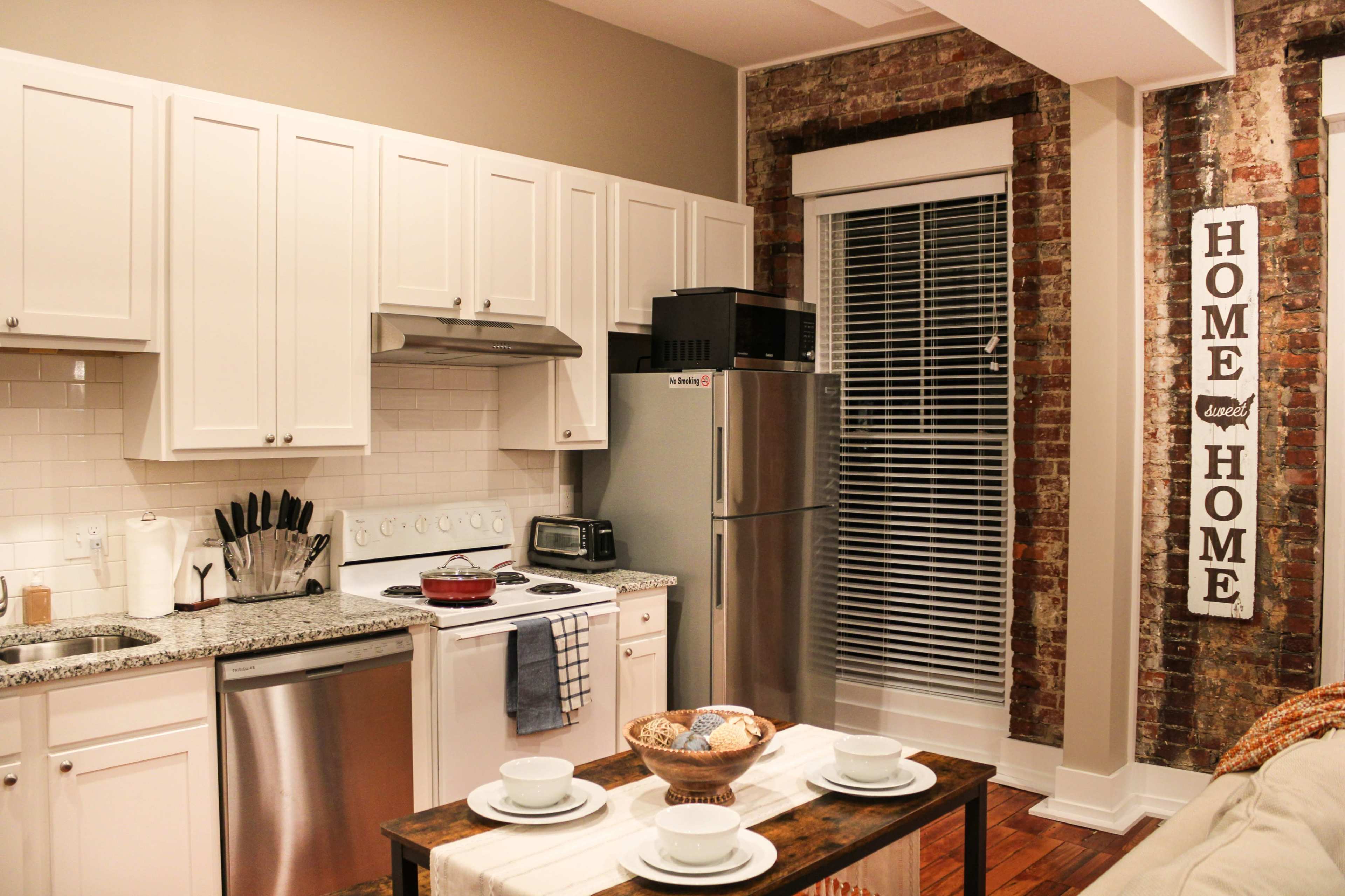 A modern kitchen features white cabinetry, a stainless steel refrigerator, and exposed brick walls.