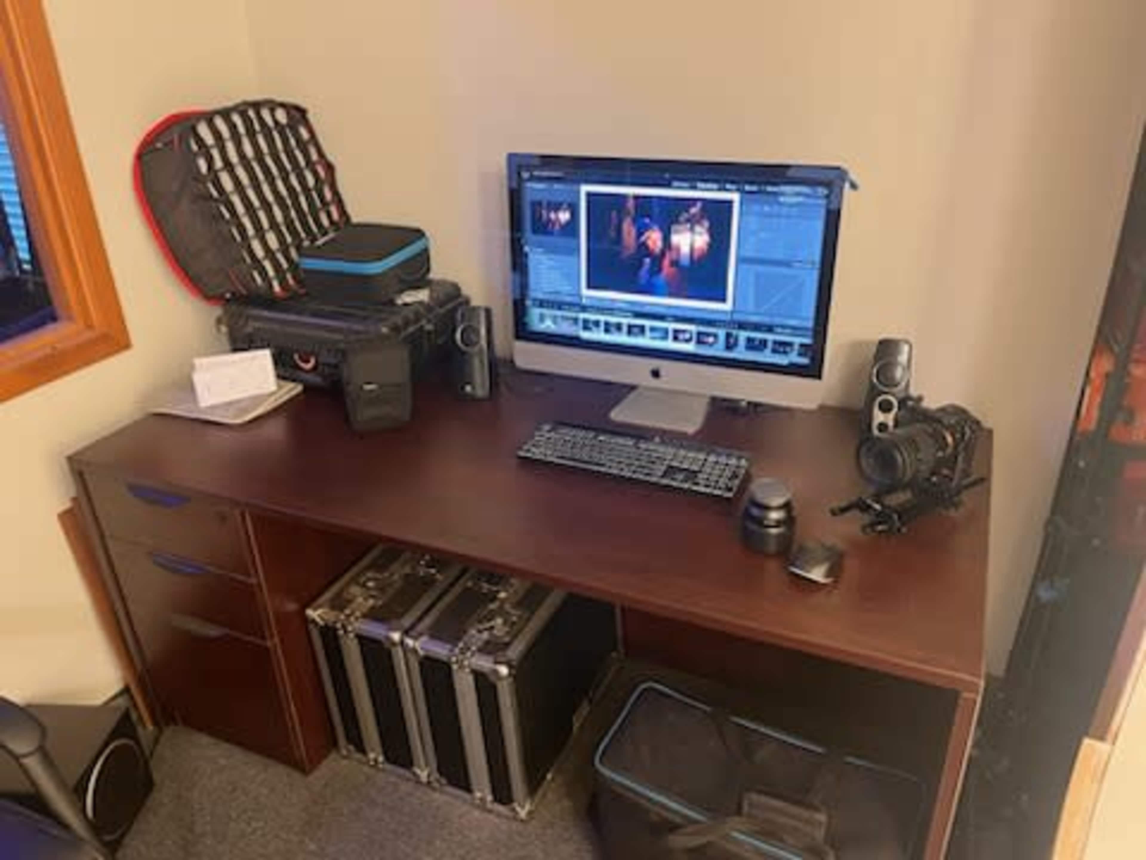 A workspace featuring a computer monitor displaying editing software, surrounded by camera equipment and storage cases on a wooden desk.