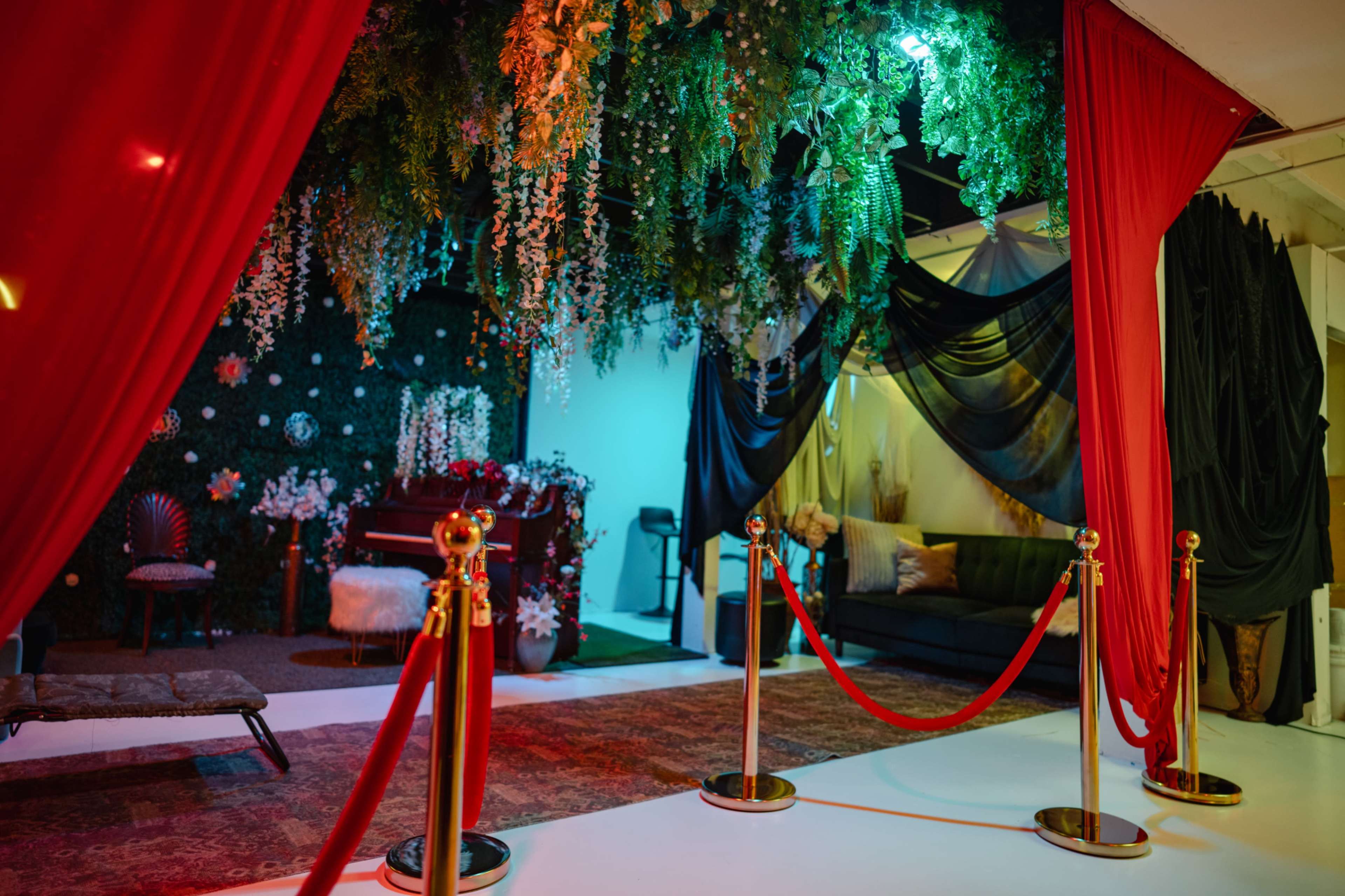 The image shows an elegantly decorated event space with draped fabric, lush greenery, and a seating area, all framed by a red carpet and velvet ropes.