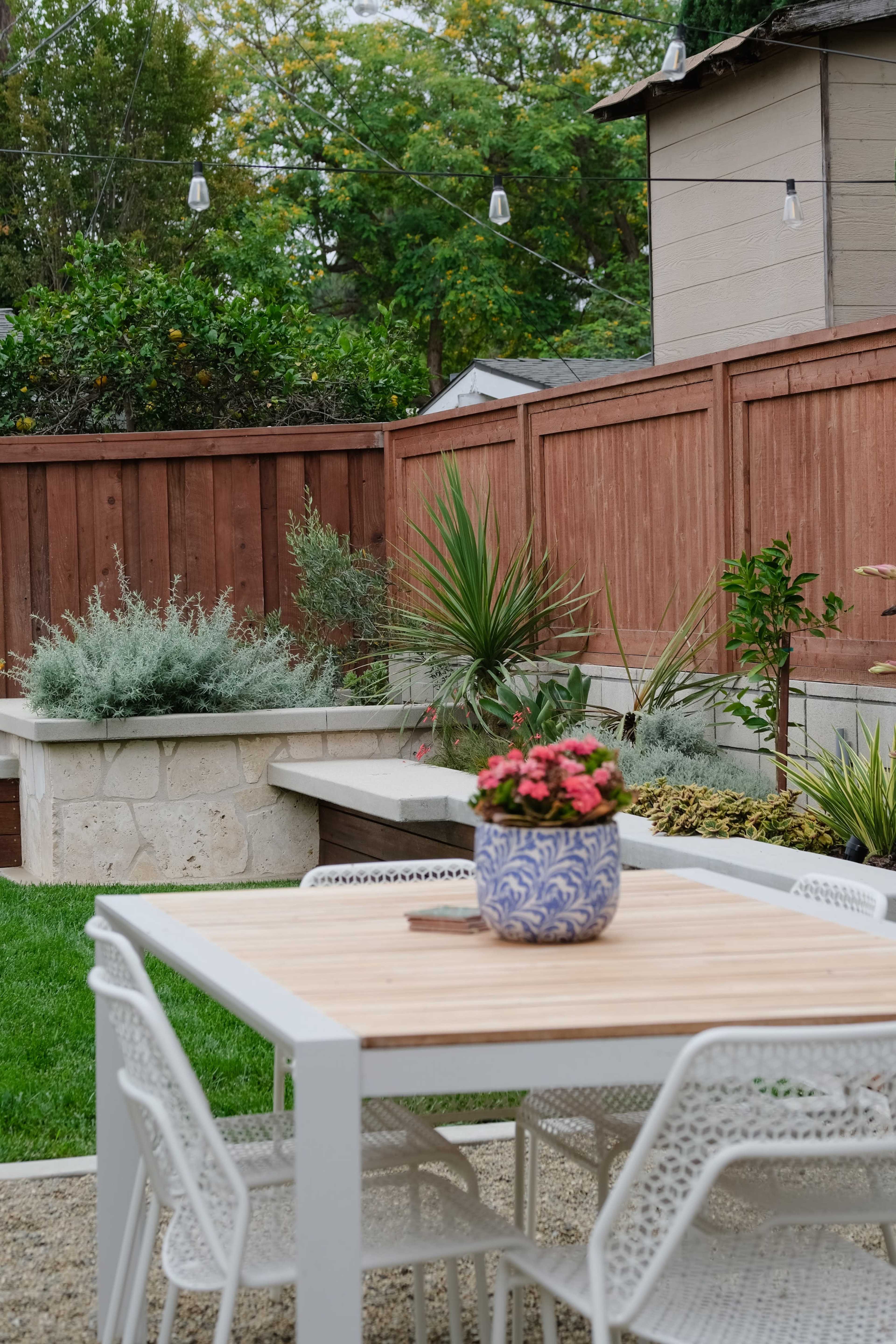 A modern outdoor dining area features a wooden table surrounded by white chairs, with a potted plant on the table and a landscaped garden in the background.