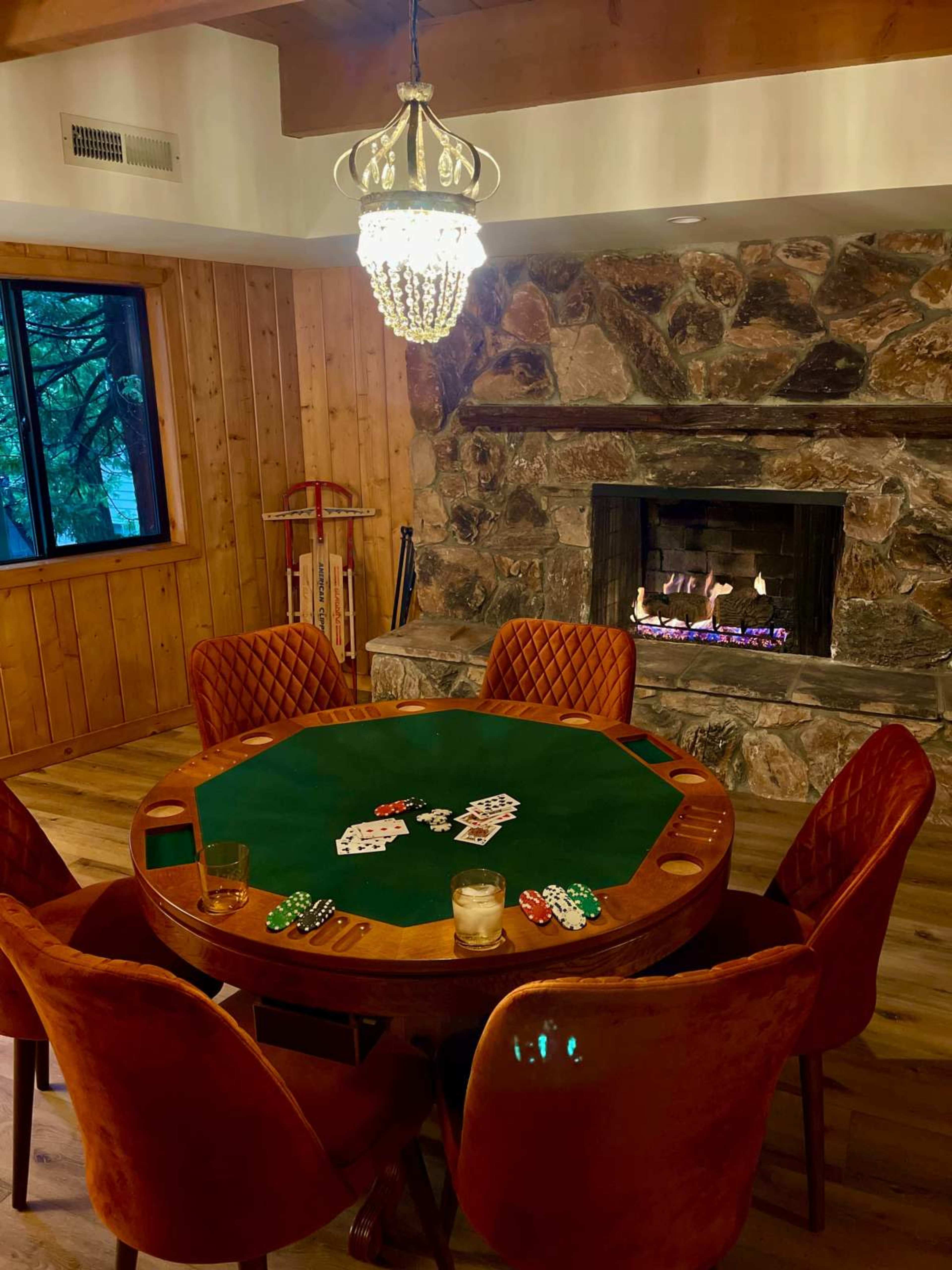 A round poker table with chips and glasses sits in a warmly-lit room featuring a stone fireplace and wooden walls.