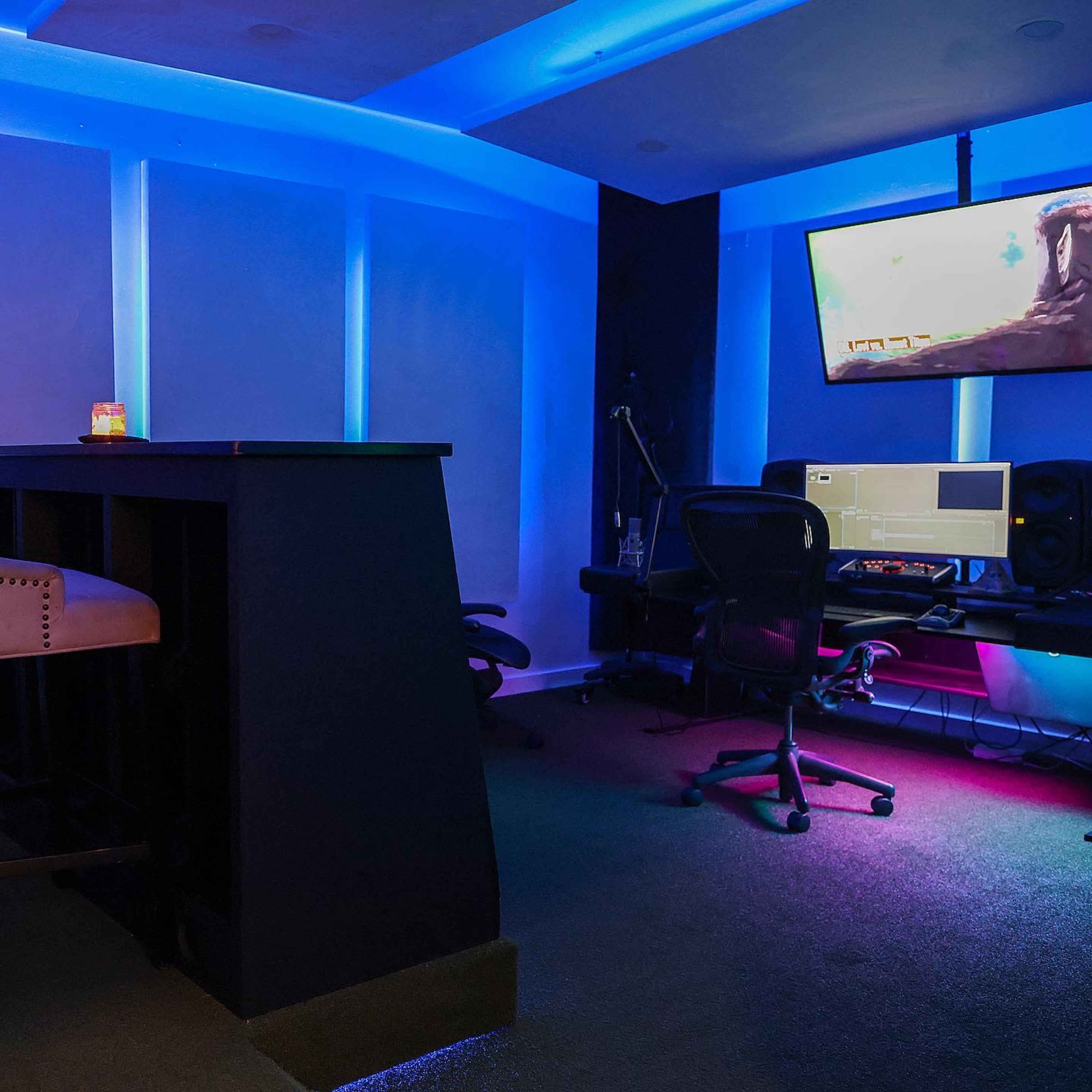 The image shows a dimly lit music studio featuring a bar area, computer workstation, and large screen with colorful lighting accents.