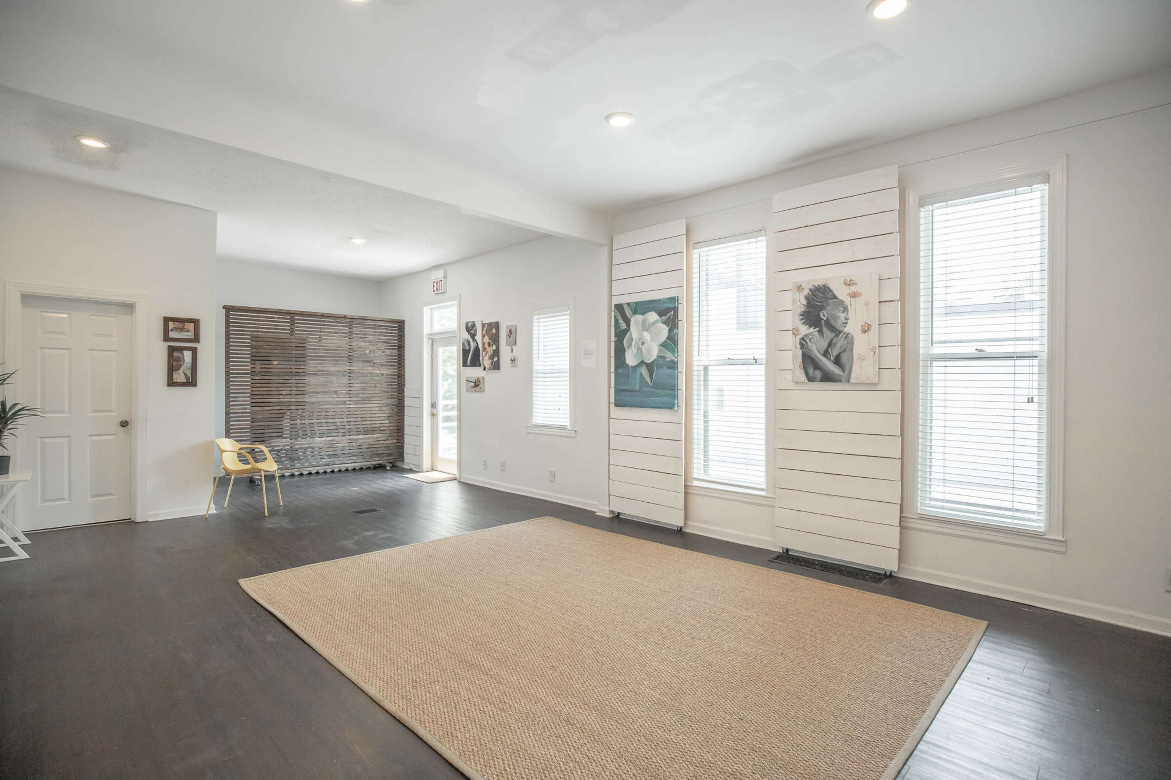 A spacious room with a large area rug, wooden floors, and natural light streaming through windows, featuring artwork on the walls and a chair in the corner.