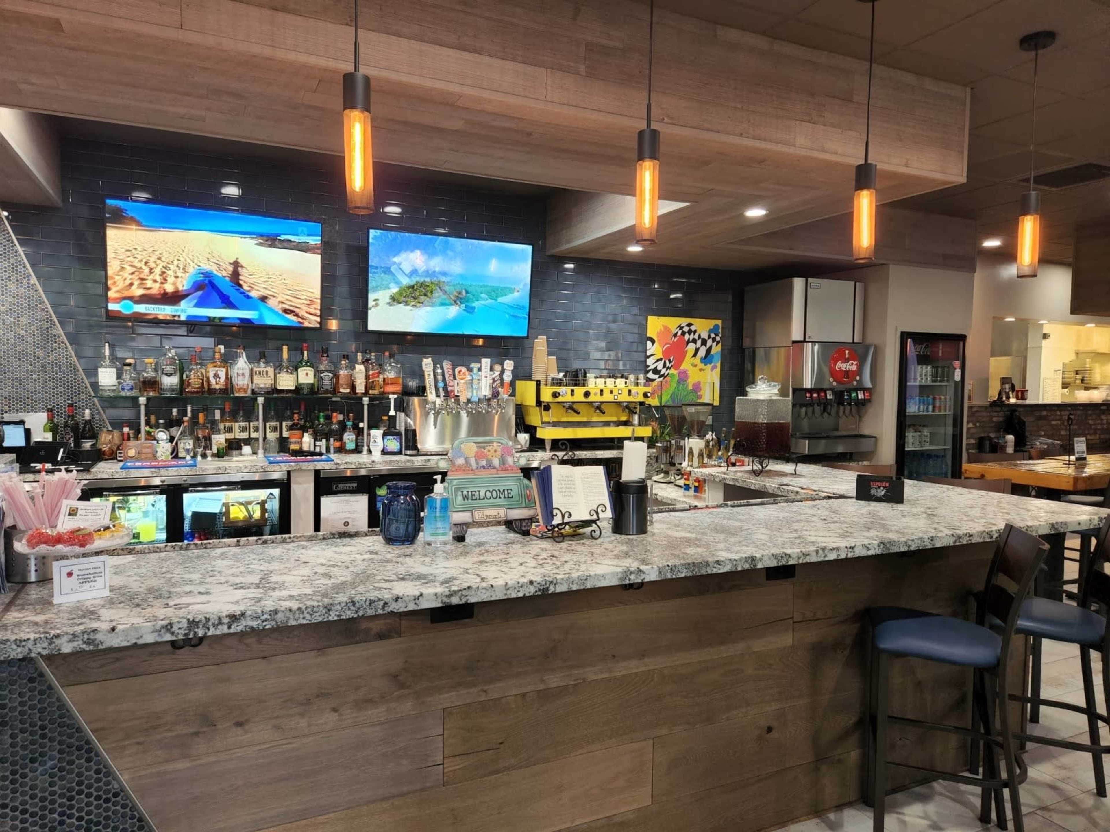 A modern bar area with two TVs displaying tropical scenes, a variety of liquor bottles behind the counter, and a menu on the bar top.
