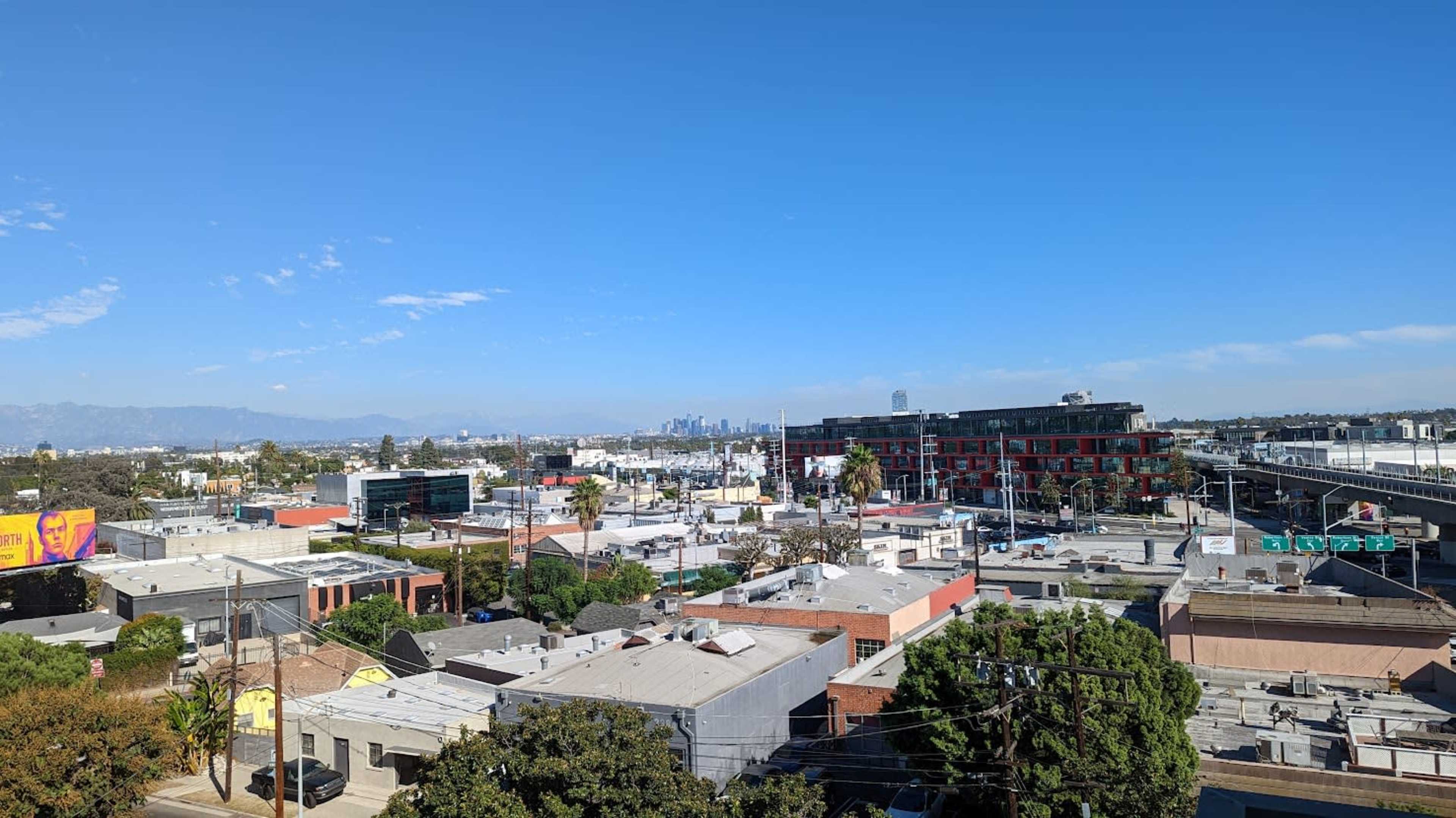 The image shows a cityscape with a view of low-rise buildings, streets, and distant mountains under a clear blue sky.