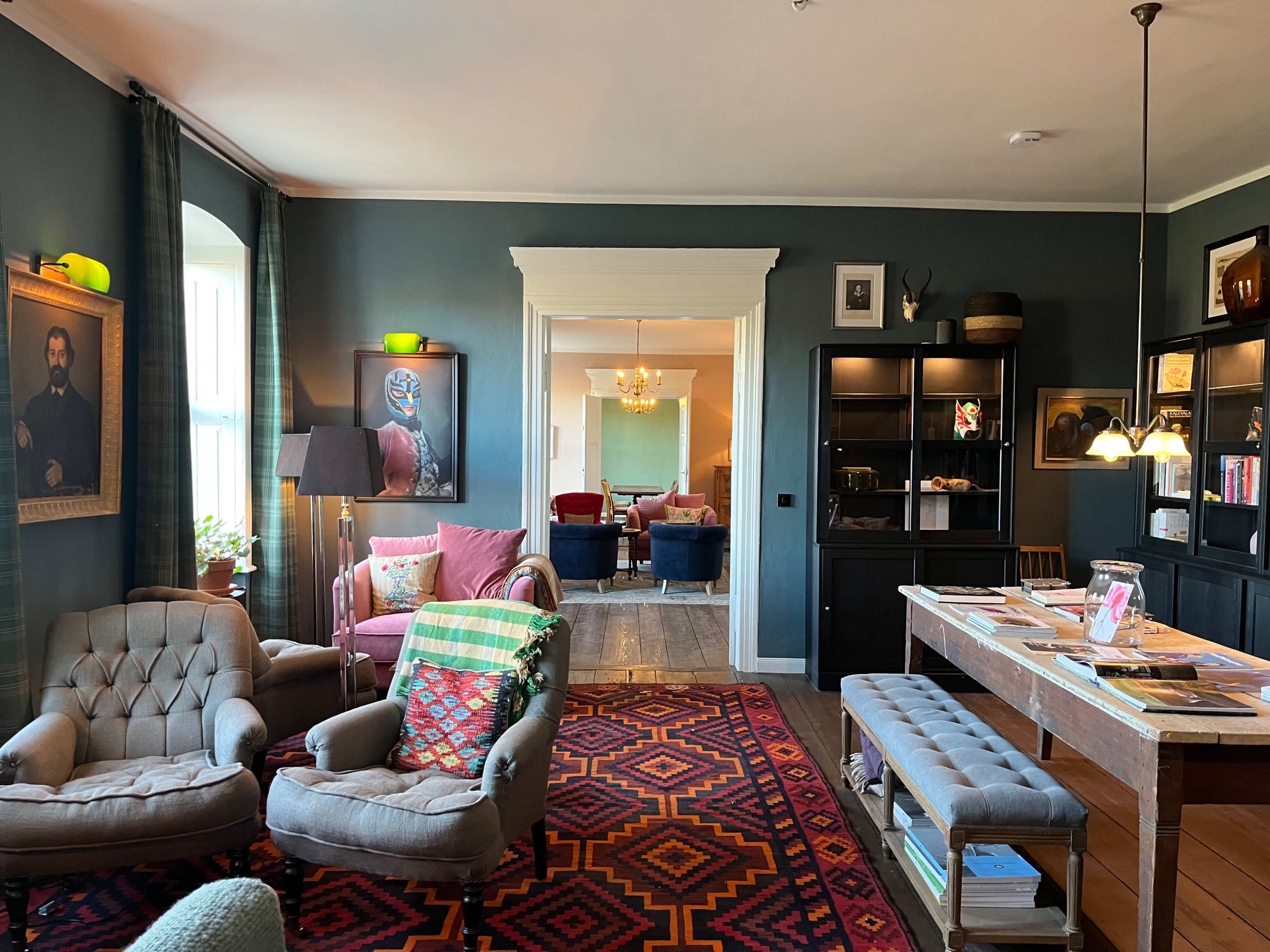 The image shows a cozy living room featuring a mix of armchairs, a wooden table, and colorful decor, with a view into another room illuminated by a chandelier.
