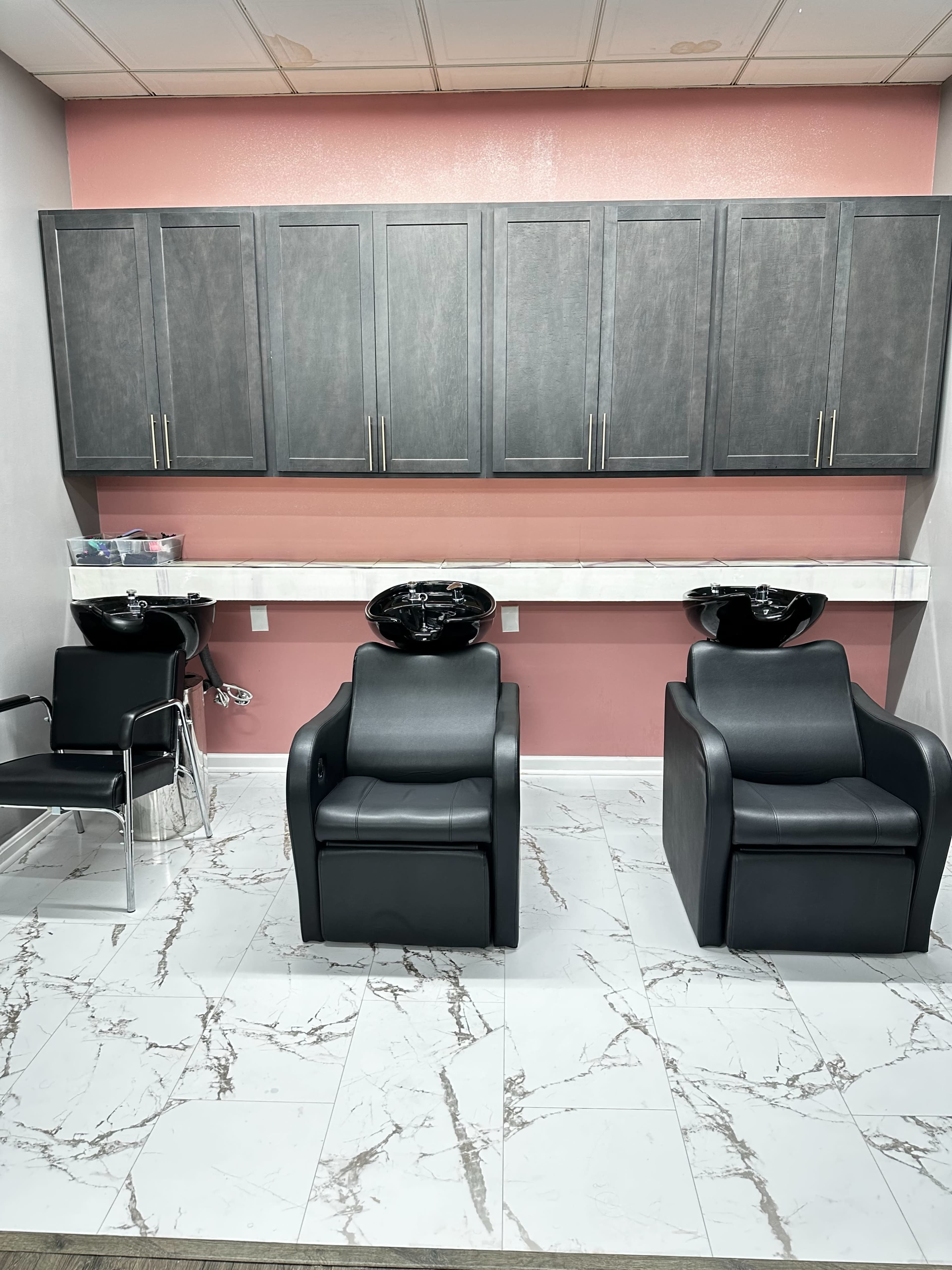 A modern salon interior with two black styling chairs and wash stations against a pink wall, complemented by gray cabinetry above and marble-patterned tile flooring.