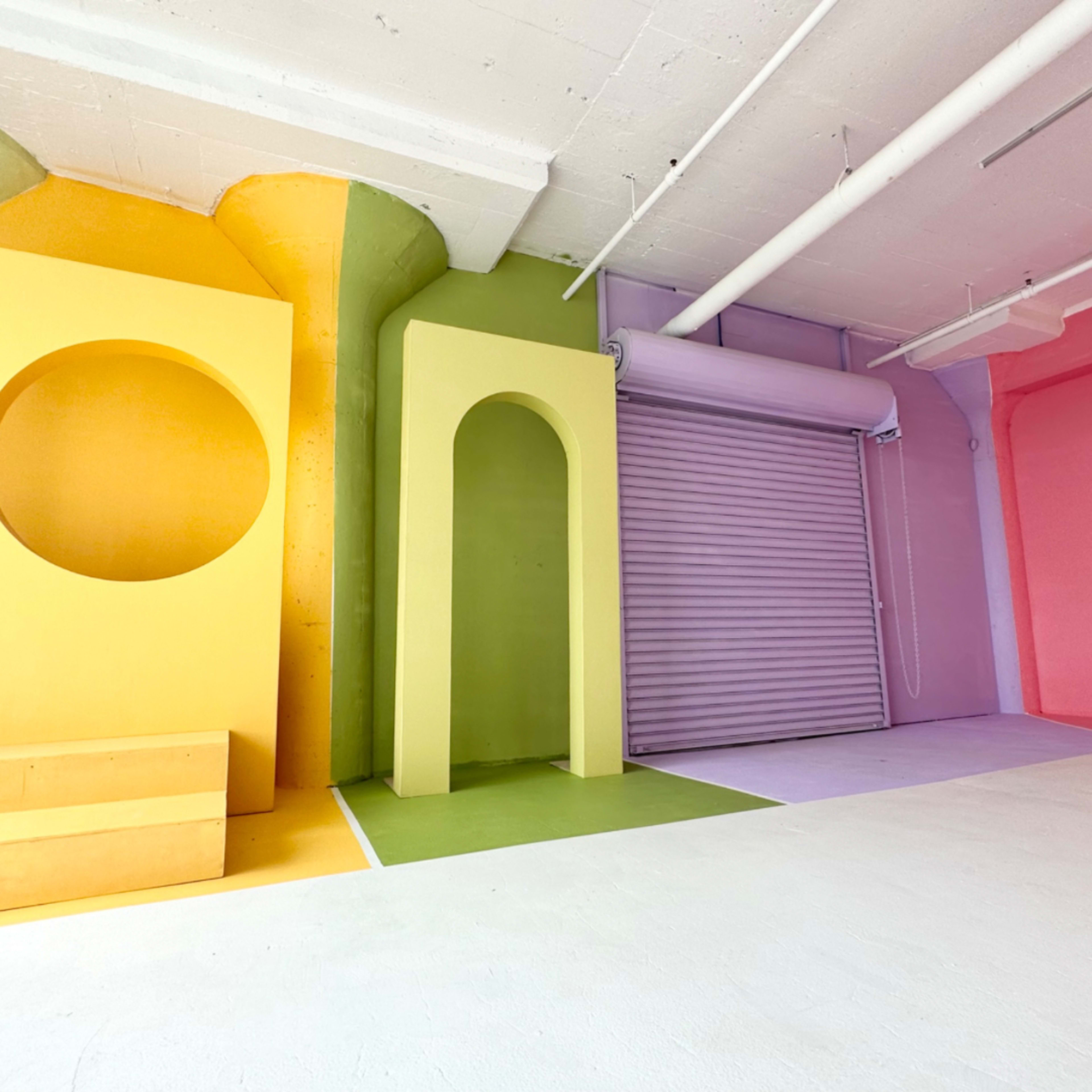 A modern interior space with brightly colored walls in yellow, green, pink, and purple, featuring geometric structures and a rolled-up door.