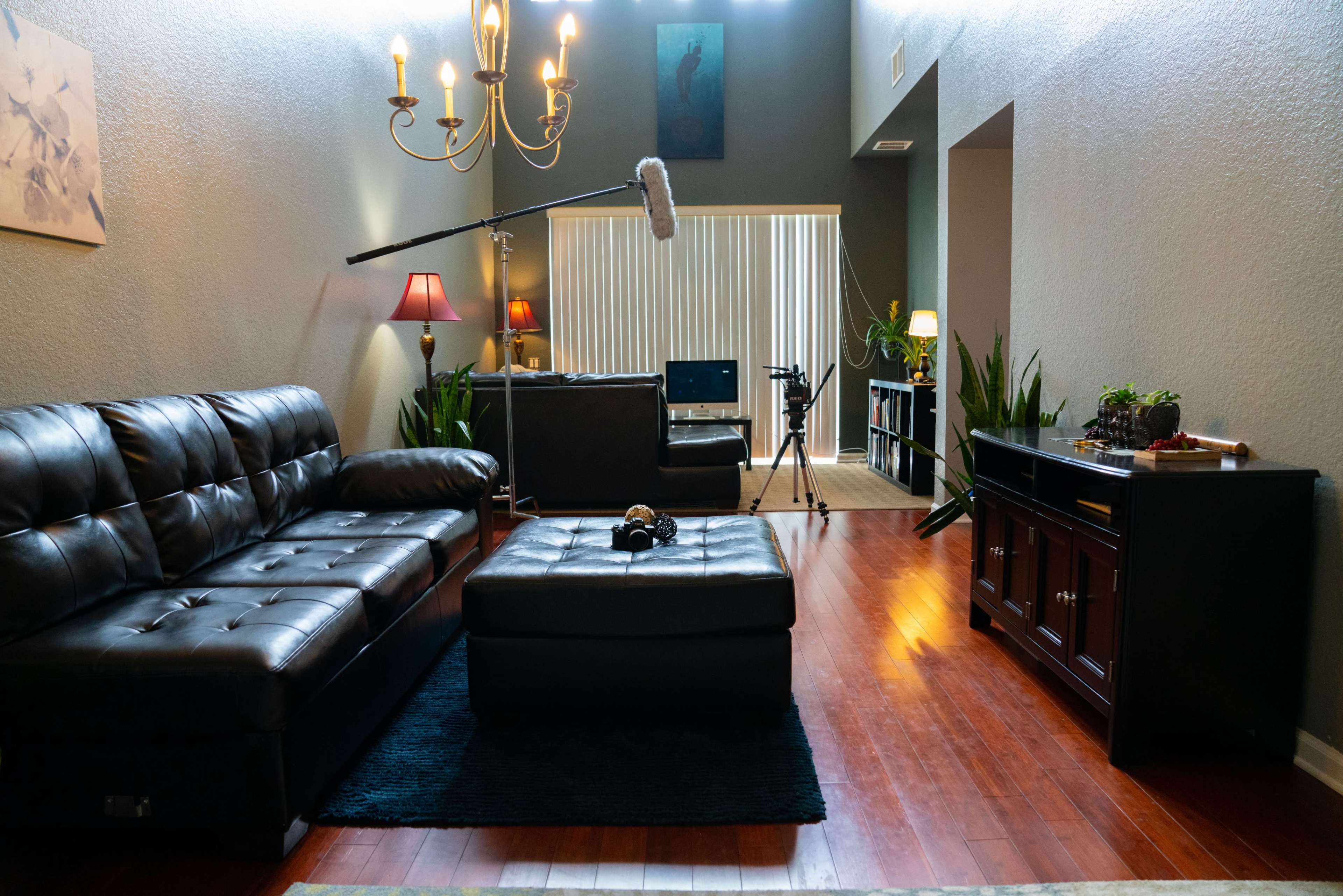 A living room with dark leather sofas, a coffee table in the center, a chandelier overhead, and a camera setup in the background.