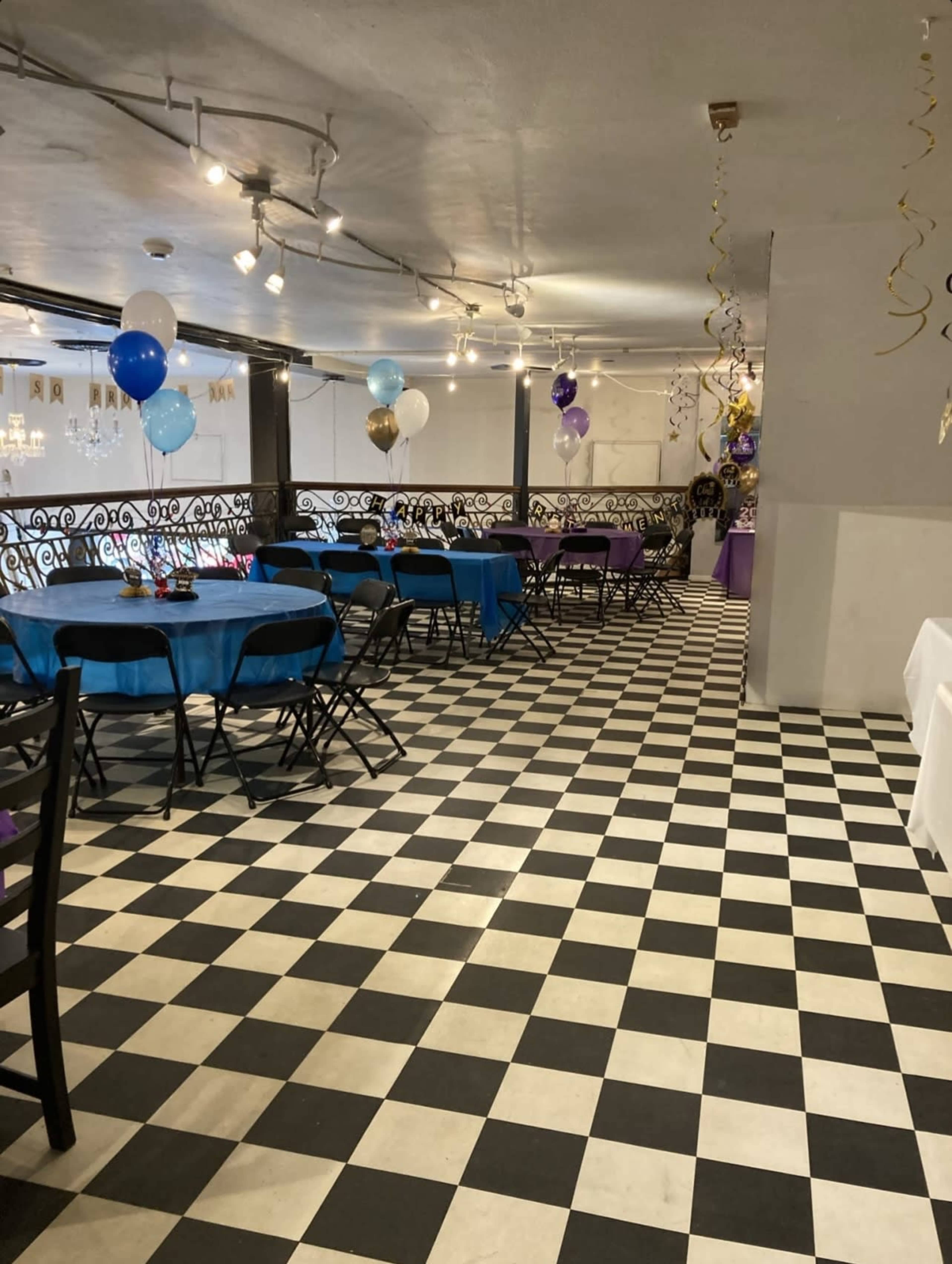 A decorated event space features black and white checkered flooring, with blue and purple balloons and tables set for a gathering.