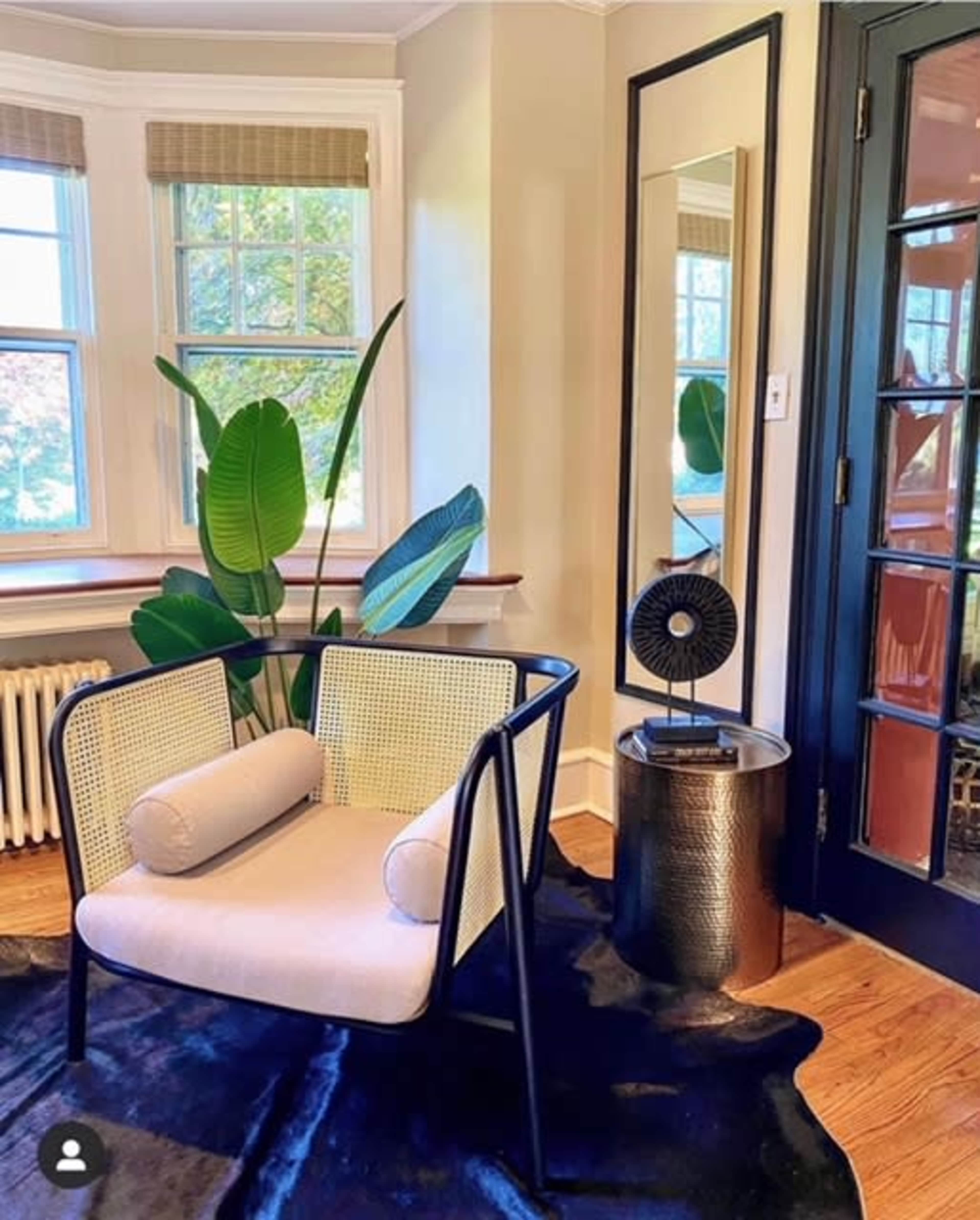 The scene features a modern chair with a circular cushion next to a round table and a small fan, set against a backdrop of large windows and a leafy plant.