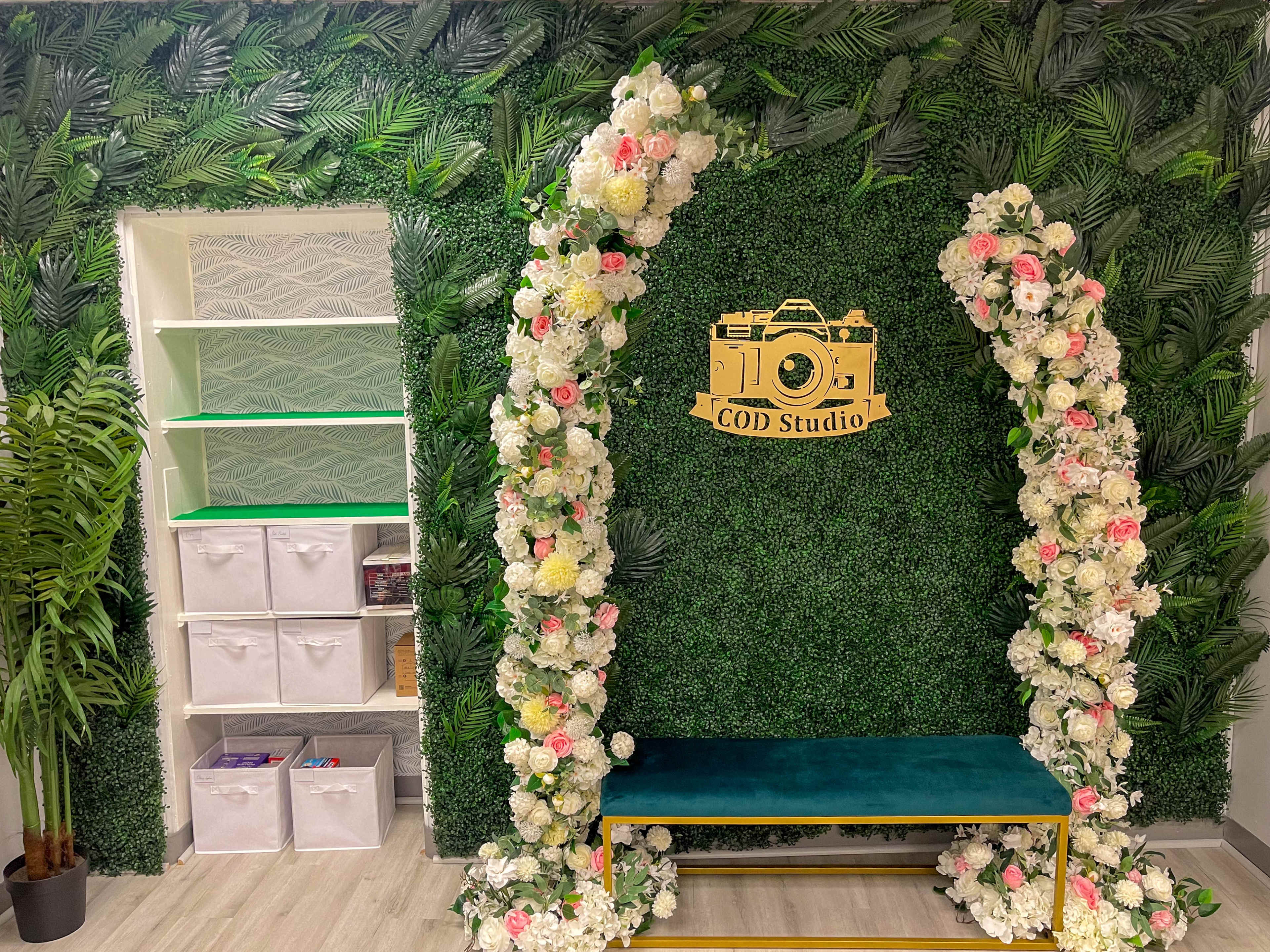 A decorated studio wall covered with faux greenery and flowers, featuring a central arch with a camera graphic and a teal bench in front.