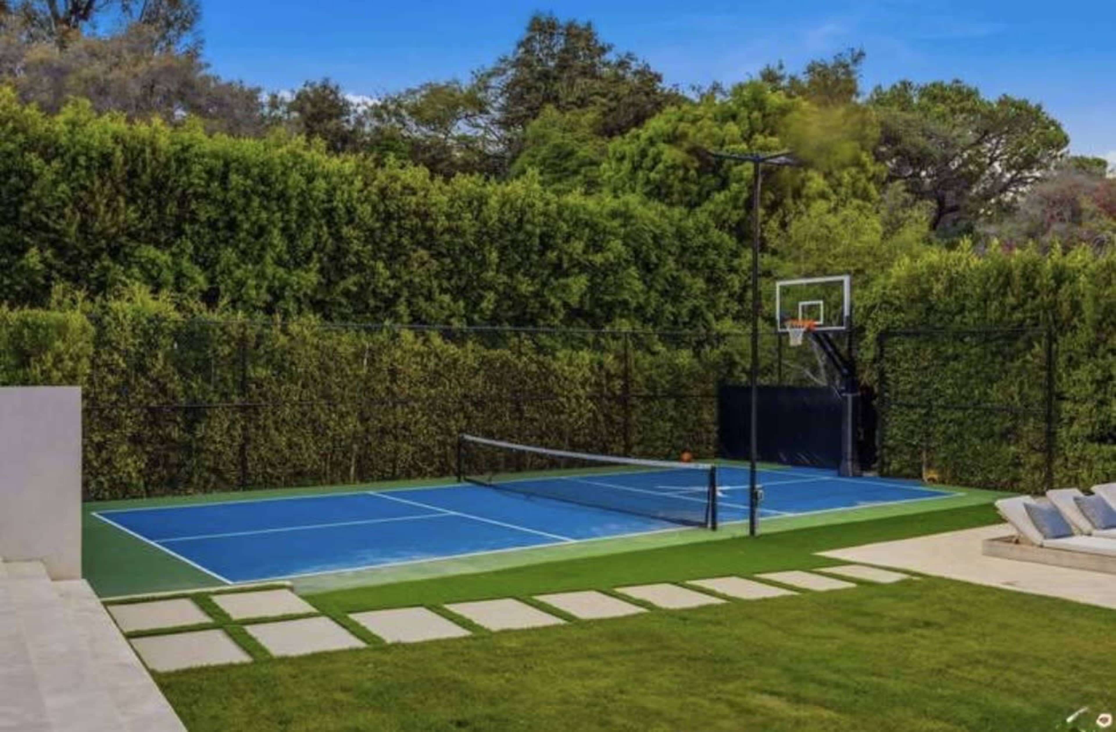 The image shows a private sports area featuring a blue tennis court and a basketball hoop surrounded by tall green hedges.