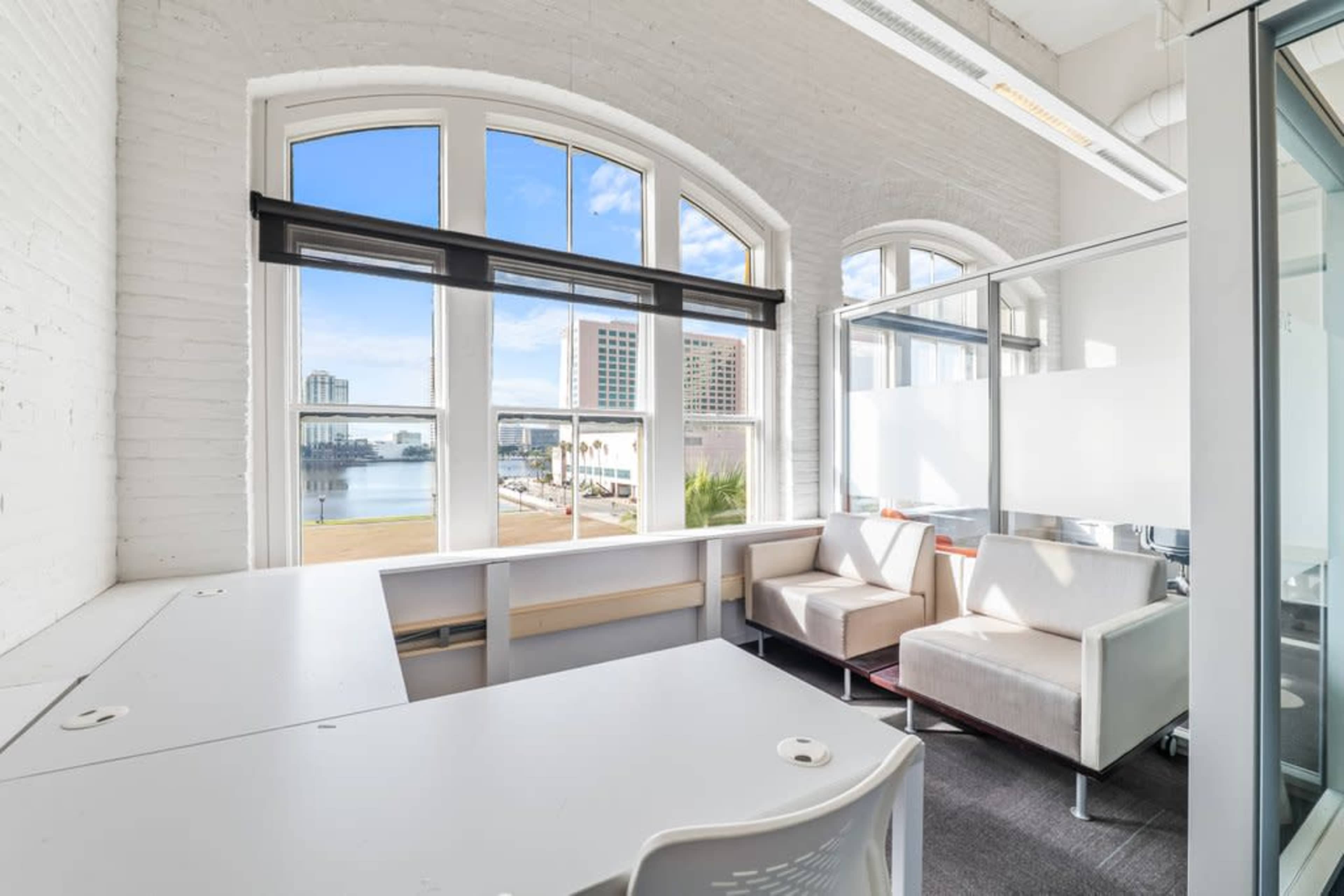 The image shows a bright office space with large windows overlooking a waterfront, featuring two white chairs and a desk along one wall.
