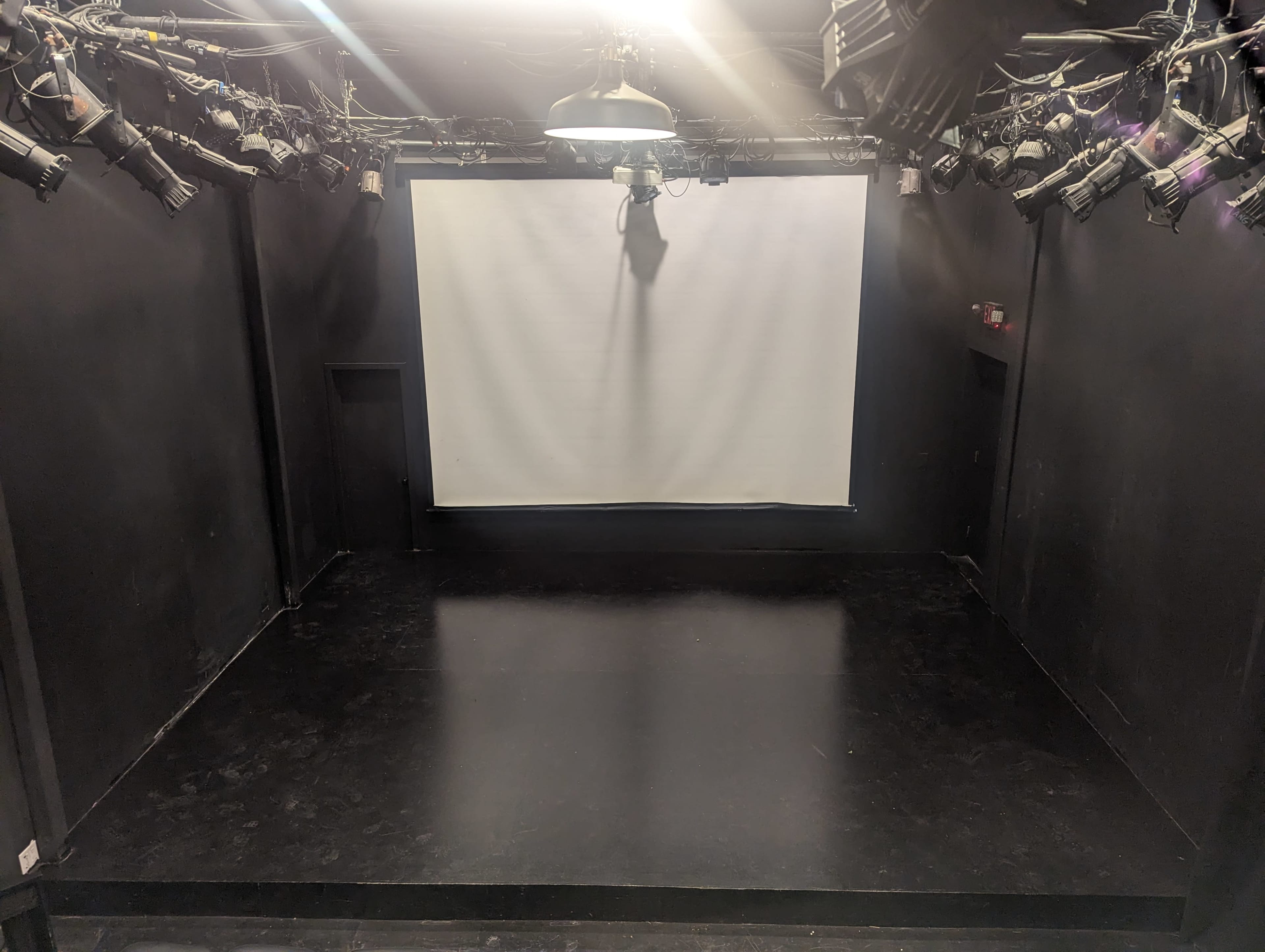 A dark theater stage features a large white projection screen at the front and ceiling-mounted lights overhead.