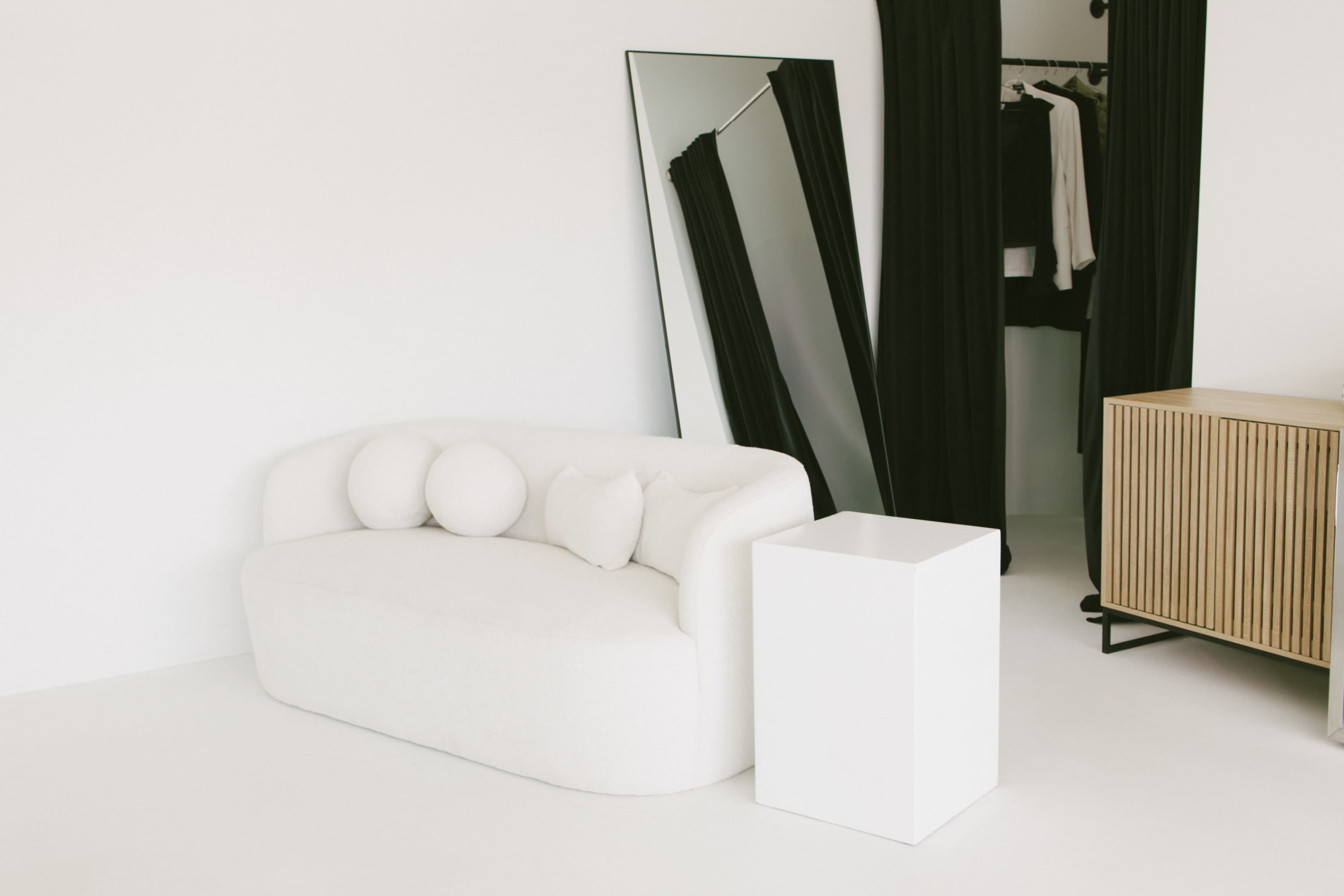 The scene features a white curved sofa with three round cushions, a large mirror against the wall, a closet with black curtains, and a wooden cabinet with a vertical slat design.