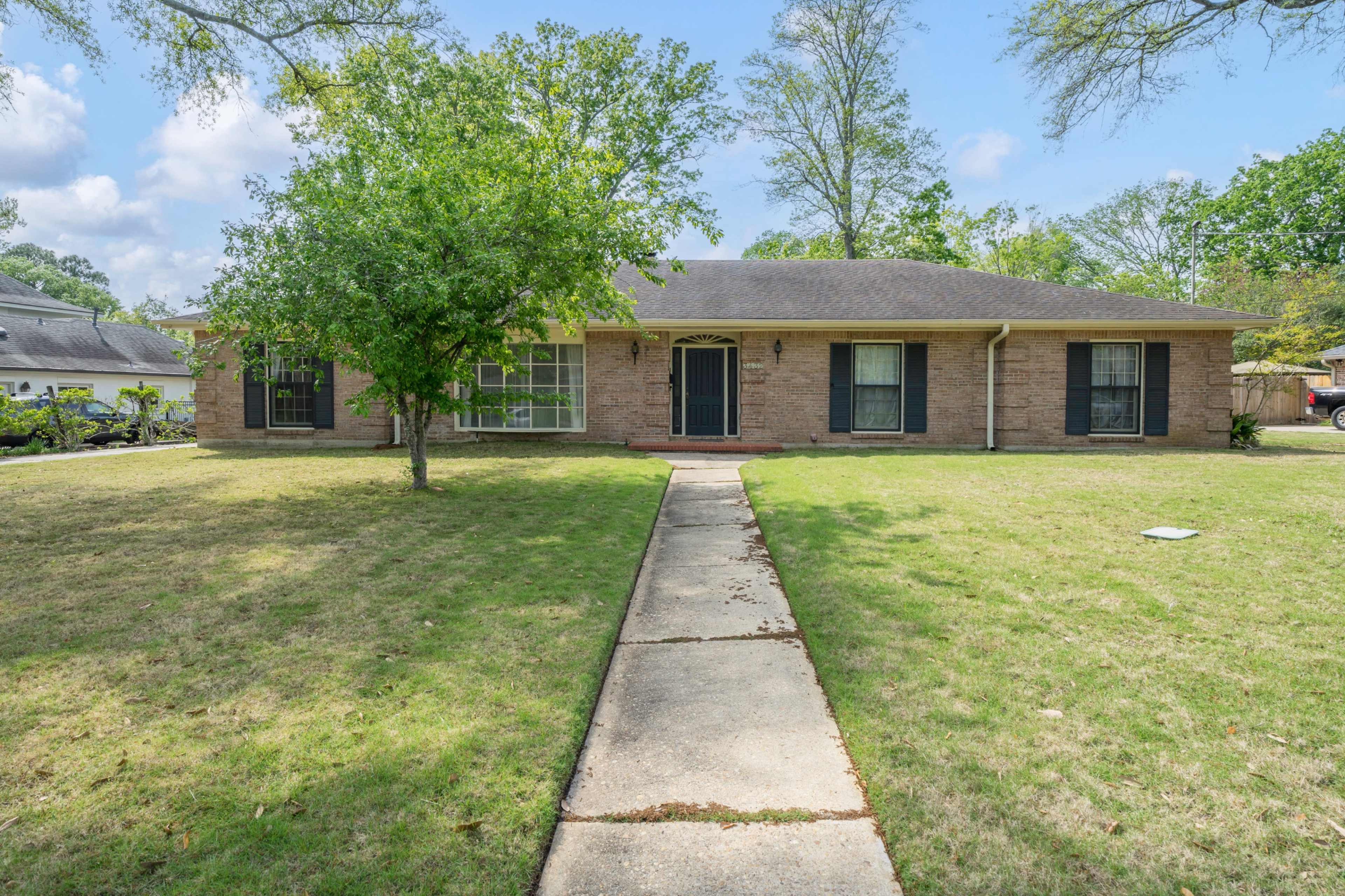The image shows a single-story, brick house with a well-maintained front yard and a concrete walkway leading to the entrance.
