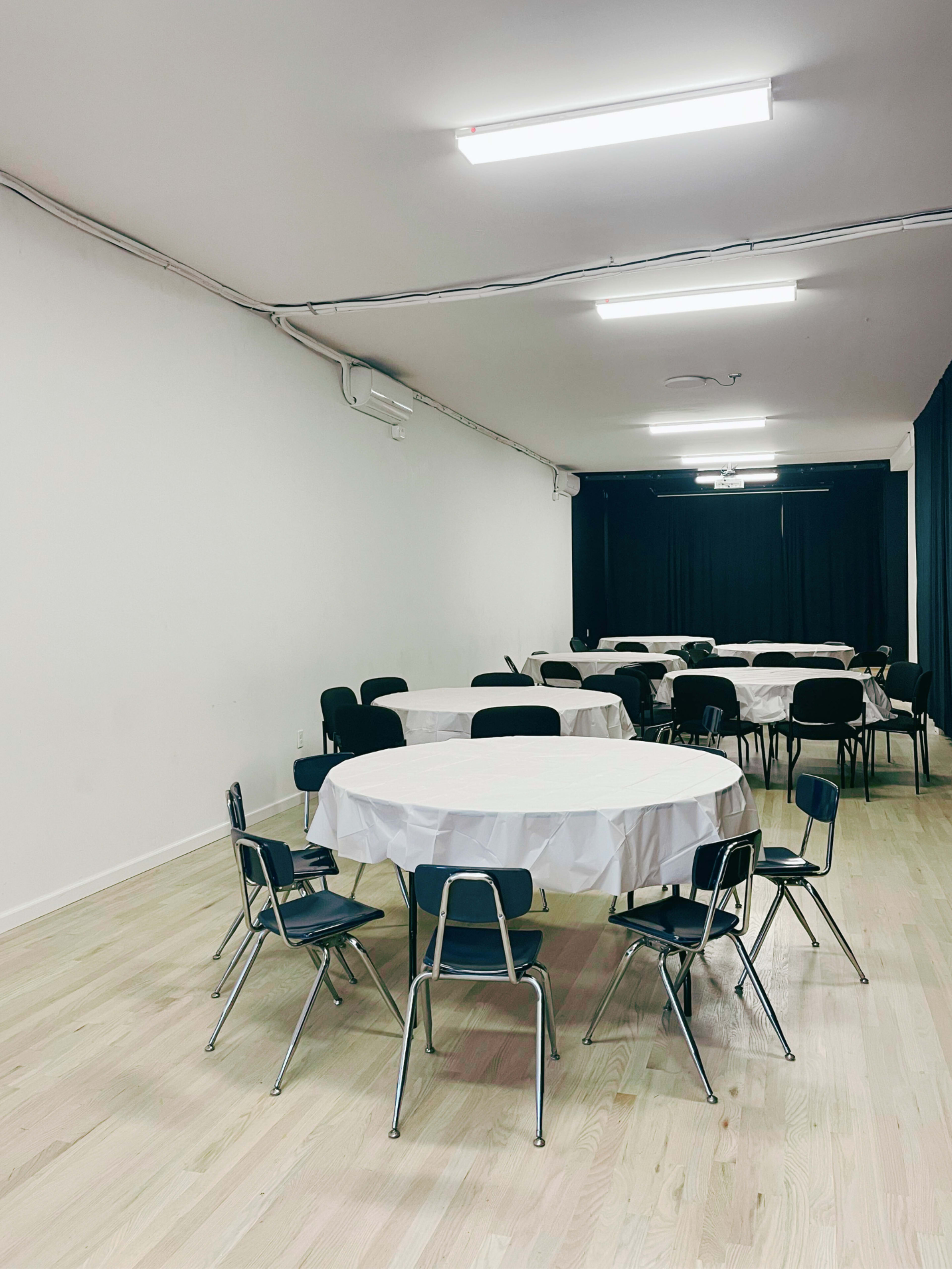 A spacious room features several round tables covered with white tablecloths, surrounded by black chairs, and has a dark curtain at one end.